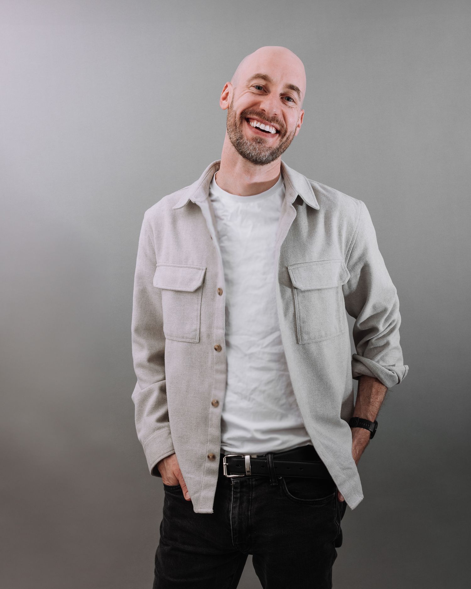 A person wearing a beige overshirt and white t-shirt stands smiling with hands in pockets against a gray background.
