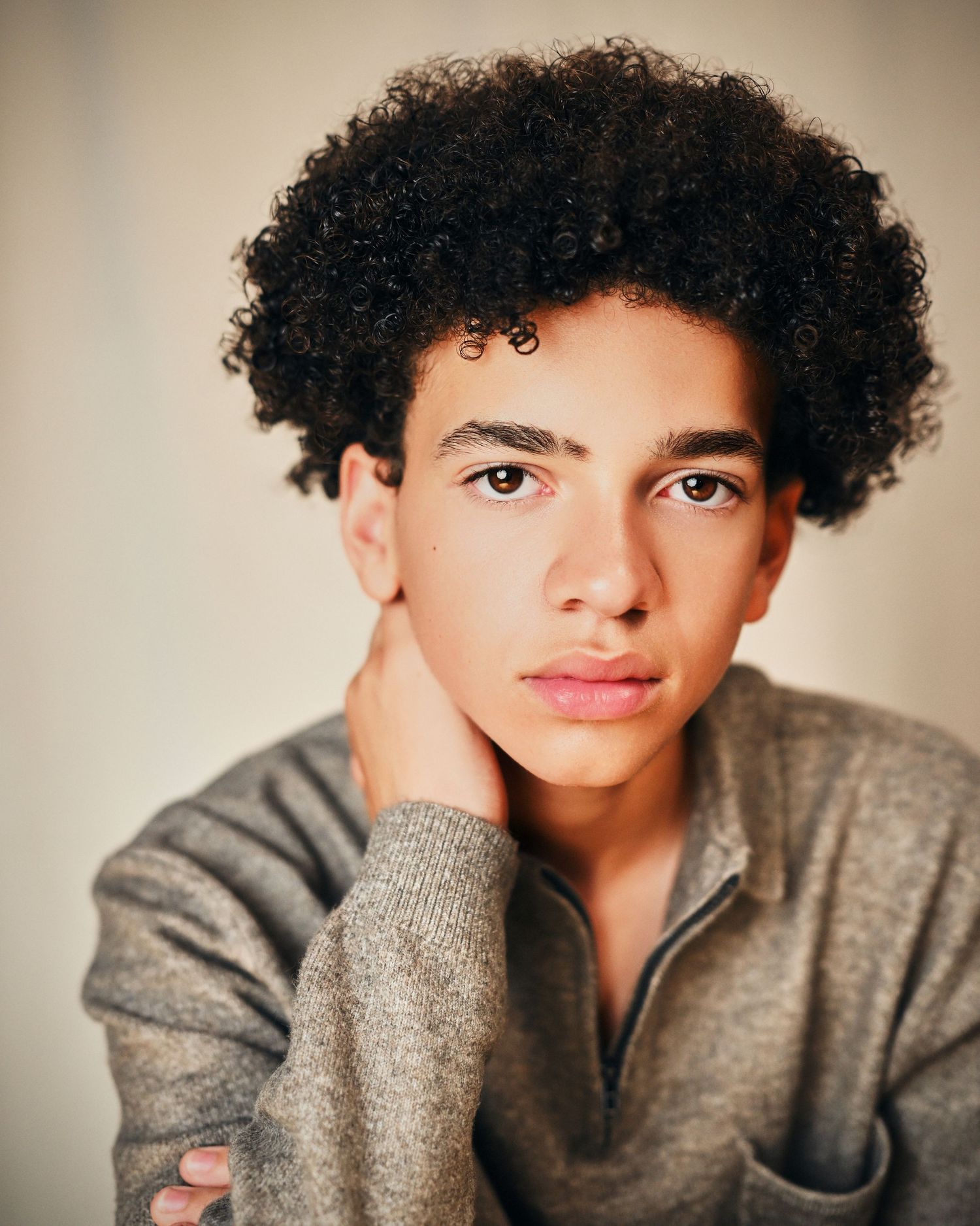 Young man with curly afro hair wearing a gray sweater, posing thoughtfully with hand on neck.
