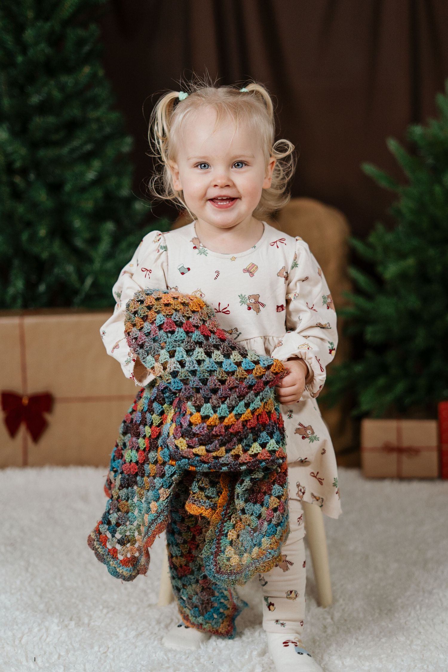 A small child in a floral outfit plays with a colorful crocheted blanket against a festive holiday backdrop.
