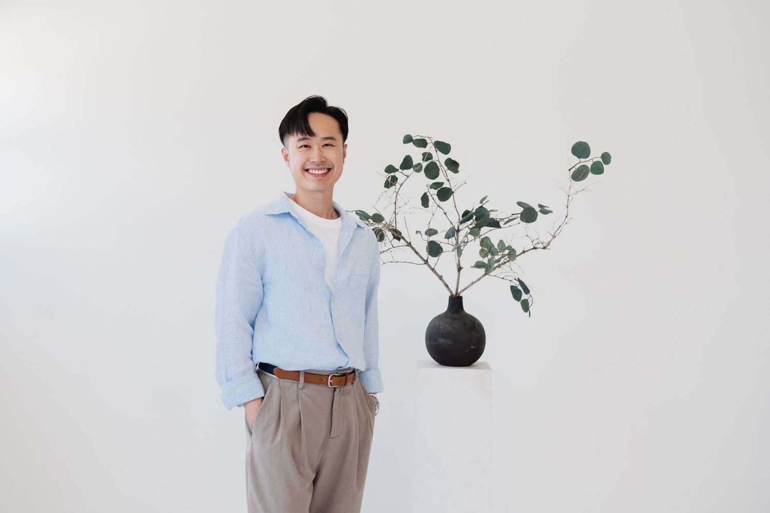 Person in light blue shirt poses next to decorative plant in black vase against white wall.