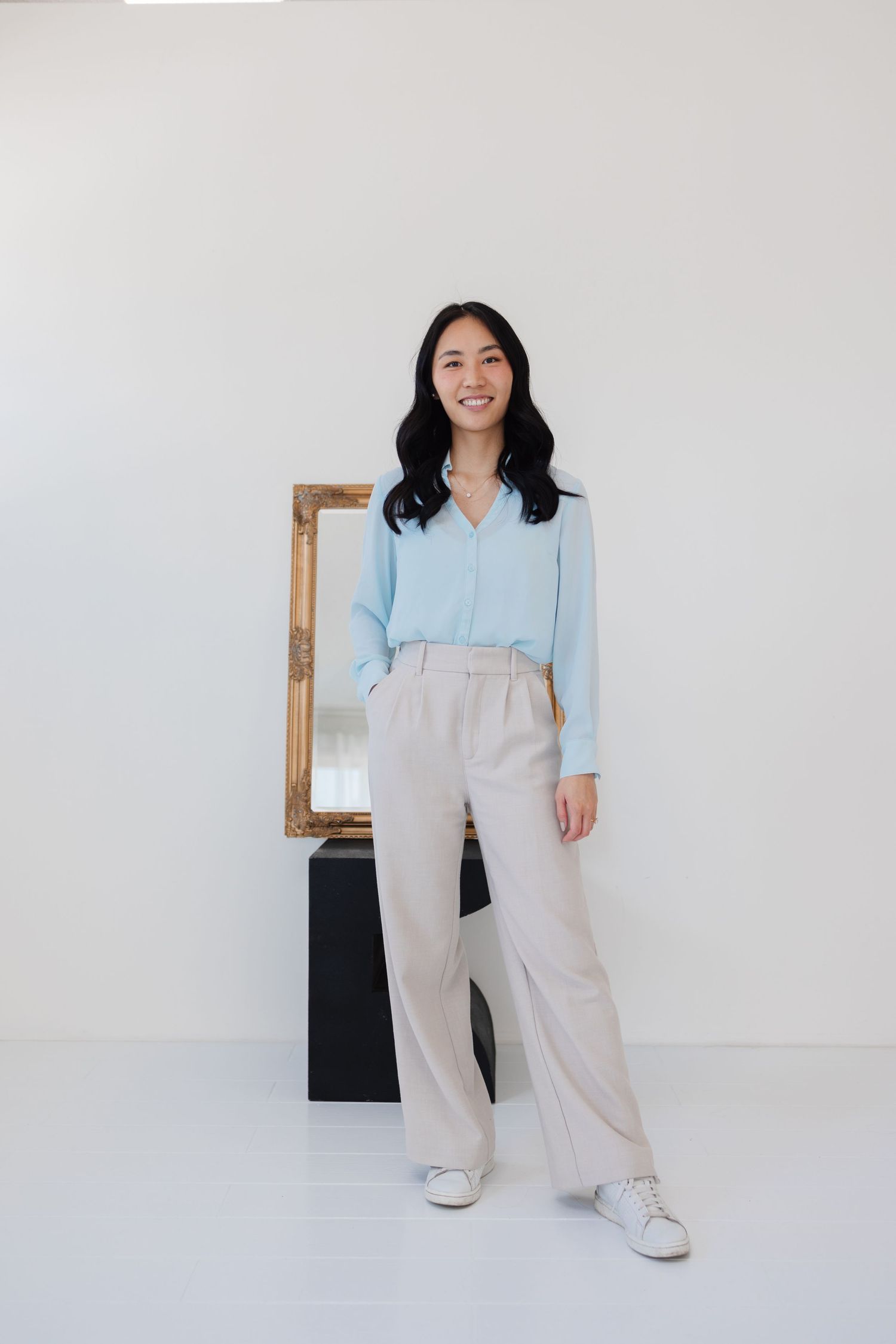 Person in light blue blouse and beige wide-leg pants stands next to vintage mirror in minimalist setting.