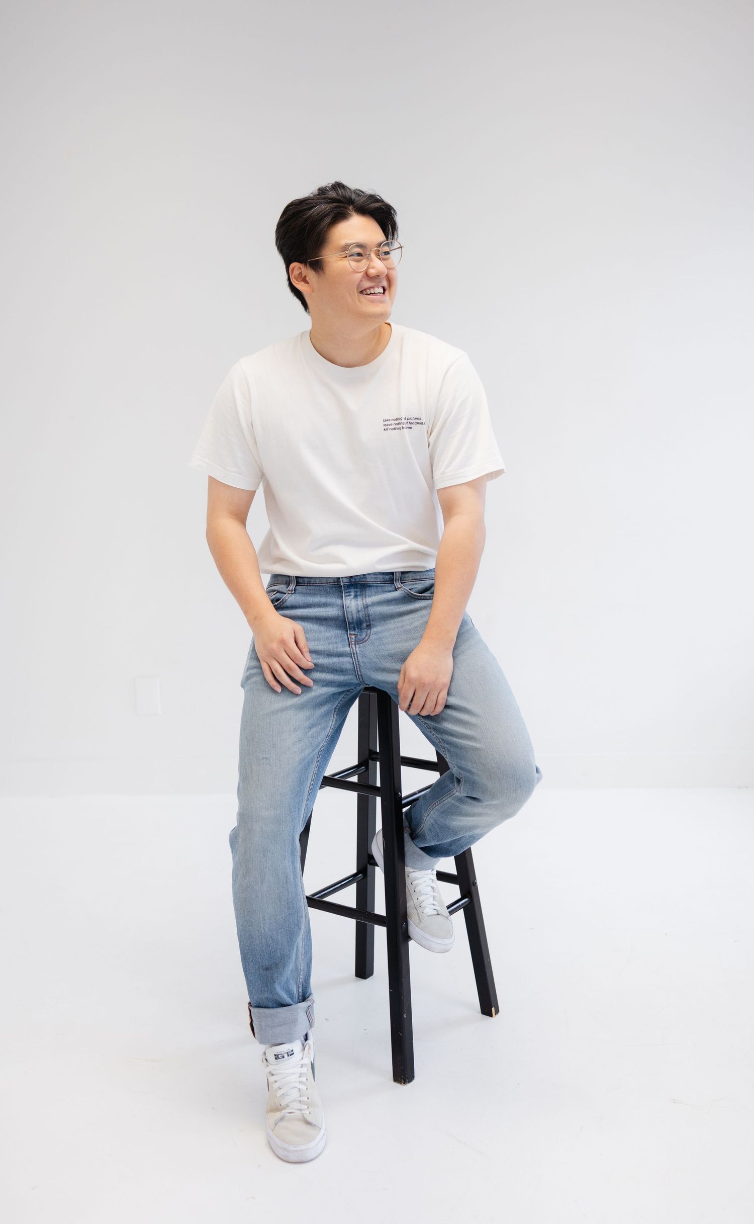 Person in white t-shirt and blue jeans sits casually on black stool against white background.