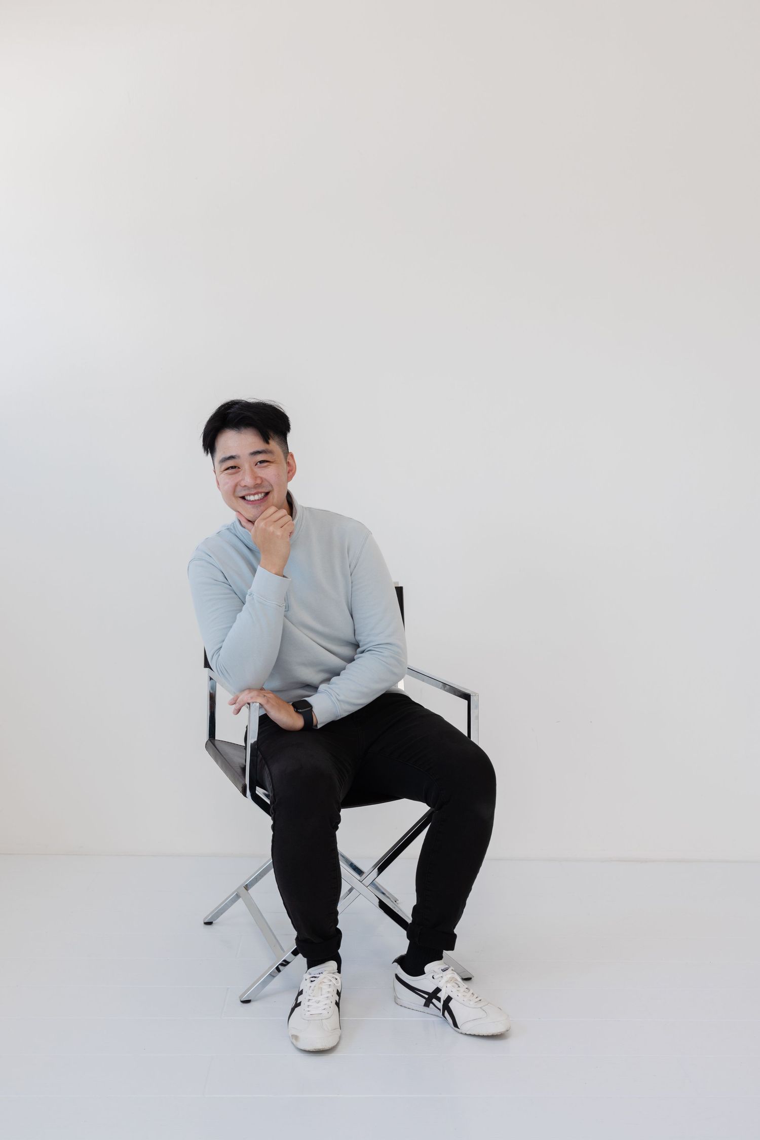 Person in gray sweater and black pants sits casually on folding chair against white backdrop.