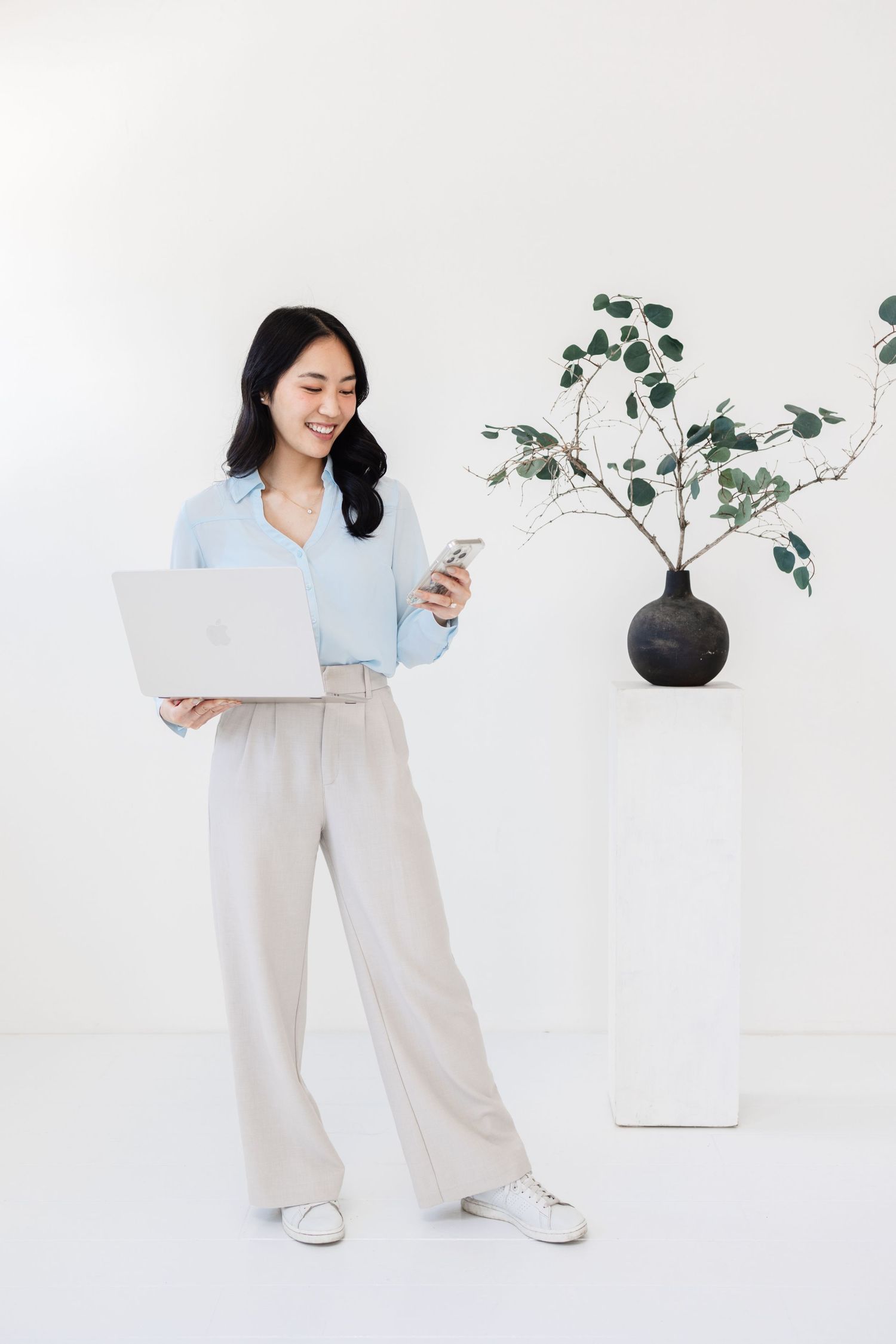 Model in casual attire using mobile device near black vase with plant against white background.