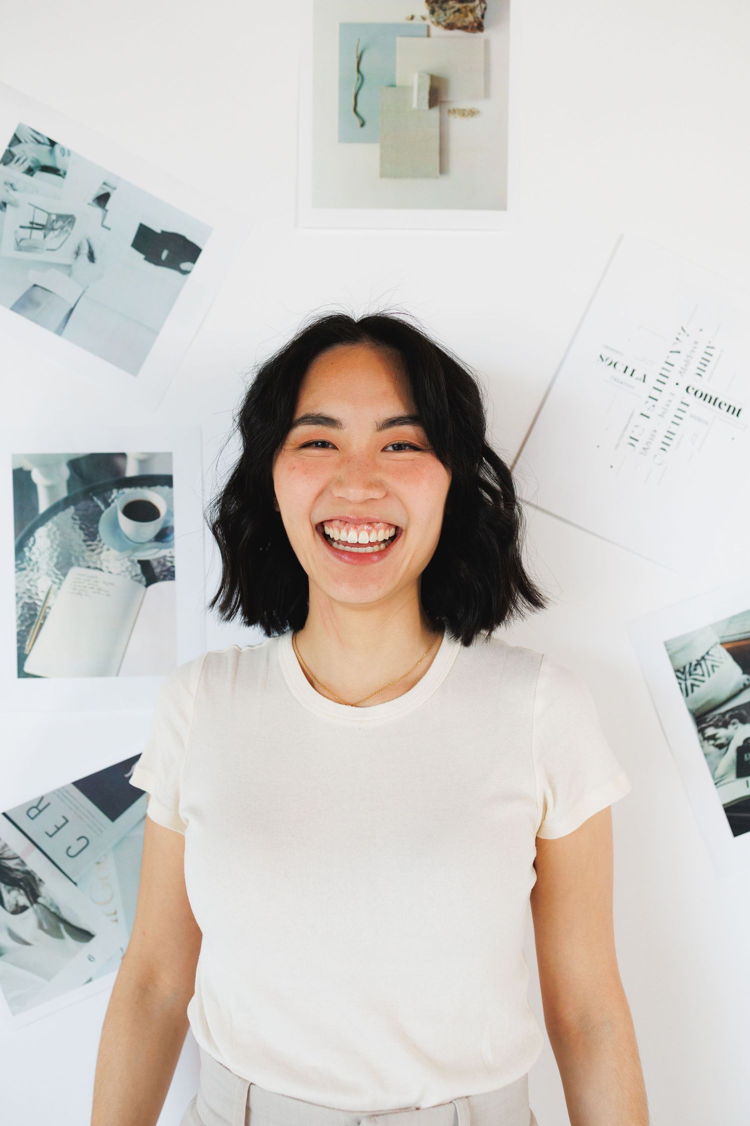 A young woman with short black hair laughs joyfully in a white t-shirt, standing before a mood board of design images.