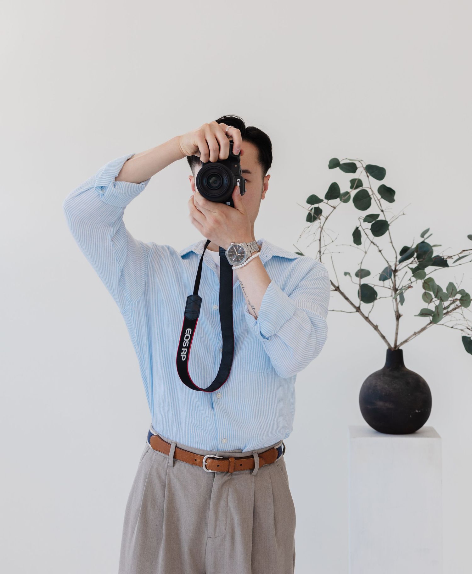 A photographer in business attire takes pictures with a DSLR camera near a black vase with greenery.