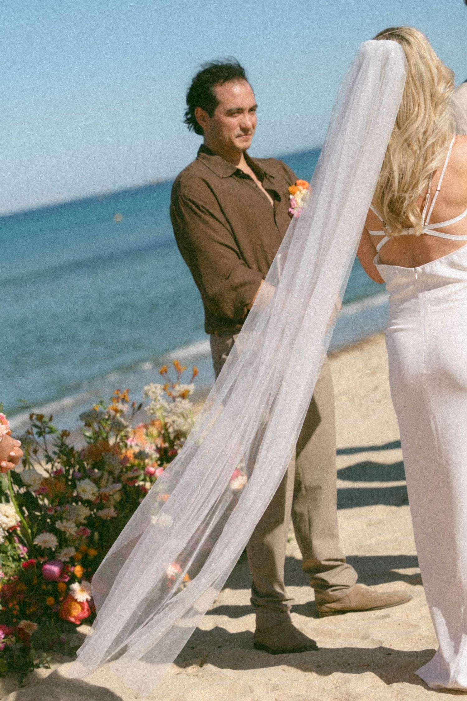A romantic beach wedding scene with flowing white veil and floral arrangements on a sandy shoreline by the ocean.