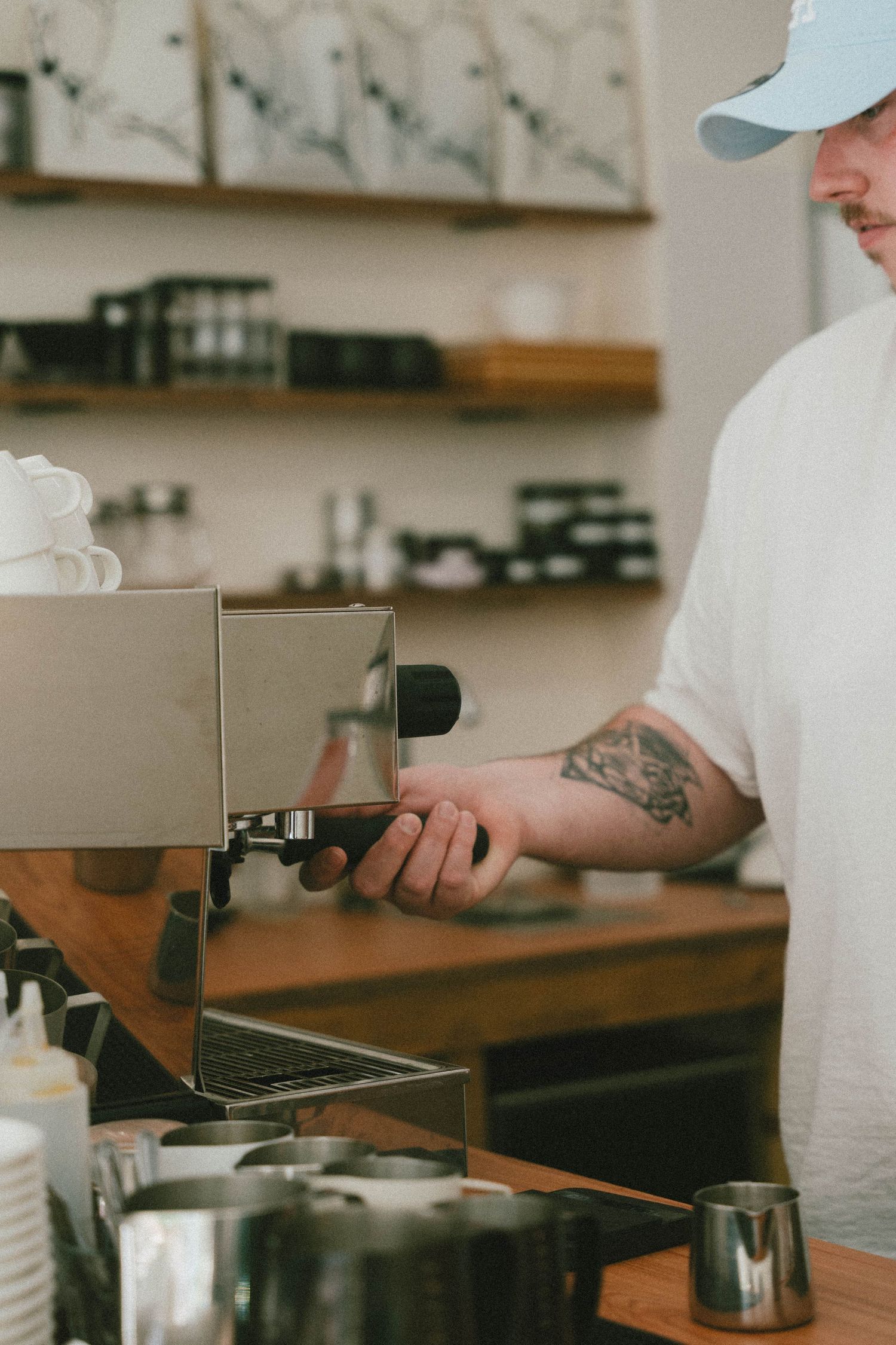 Barista tatoué en t-shirt blanc et casquette bleue utilisant une machine à expresso dans un bar à café en bois.
