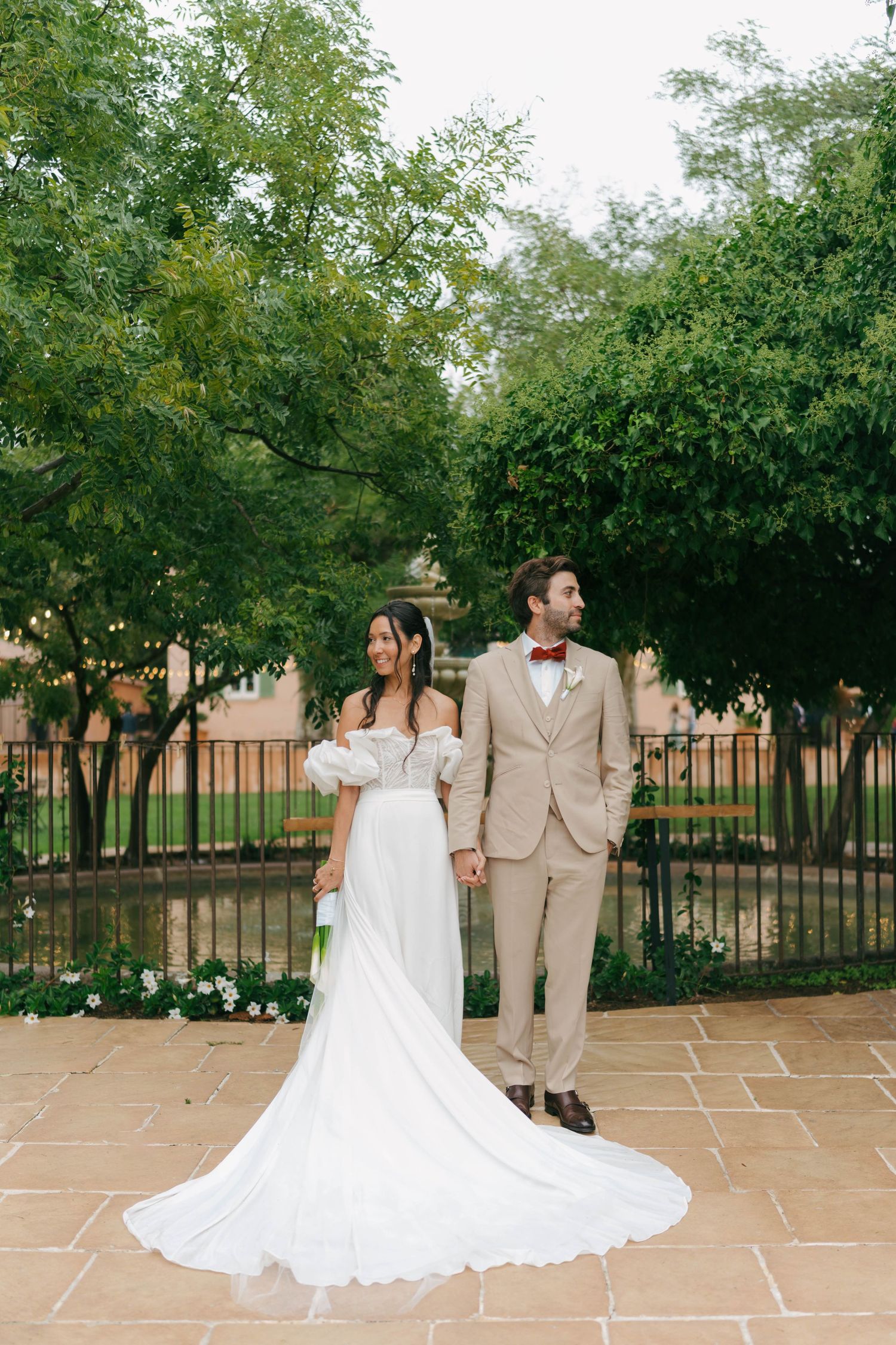 Un couple en tenue de mariage partage un moment romantique dans une cour de jardin avec des pavés en pierre et des arbres verts.