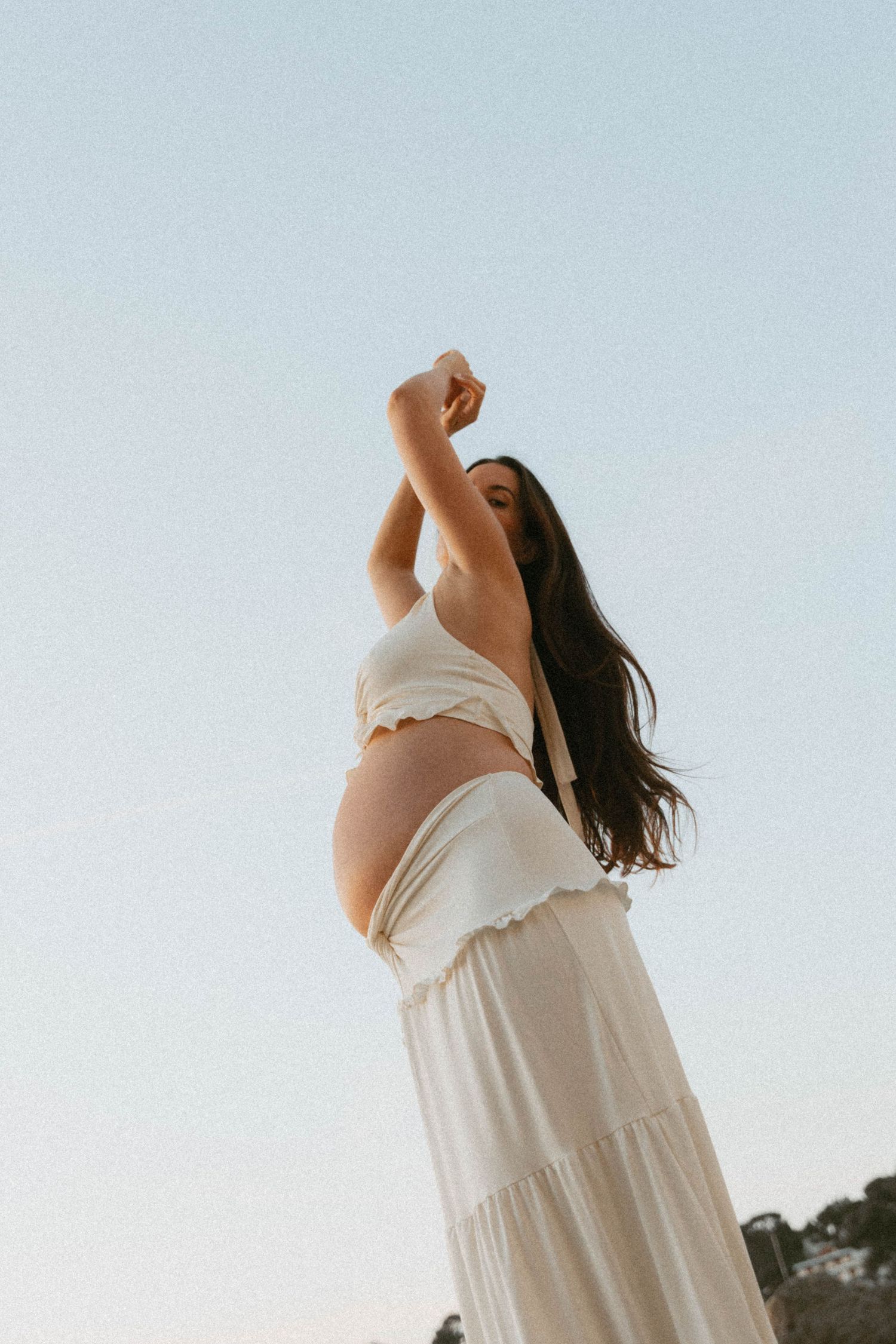 Pregnant woman in white crop top and maxi skirt poses with arm raised against a clear blue sky.