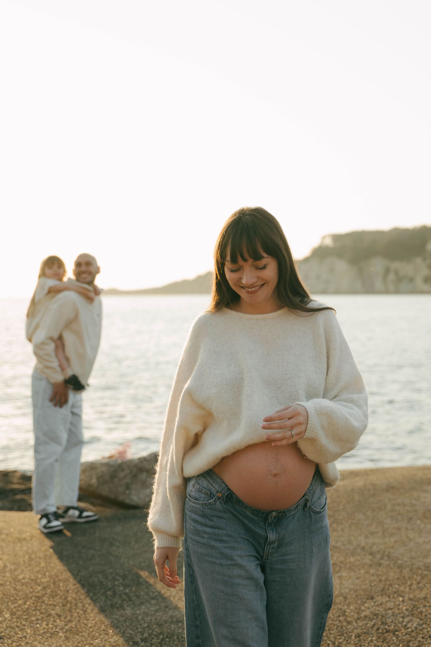 Série de photos de maternité montrant une femme enceinte en pull crème et jean sur fond de plage au coucher du soleil.