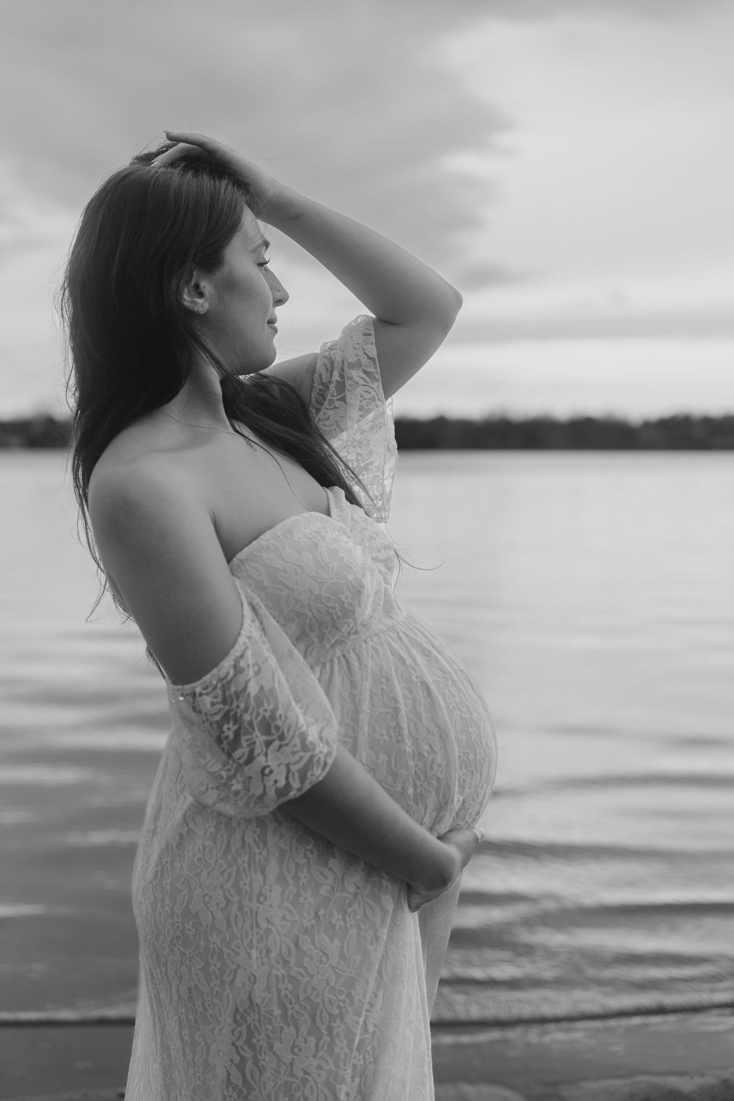 Black and white maternity photoshoot at a lake shows a woman in a lace dress posing against rippling water.