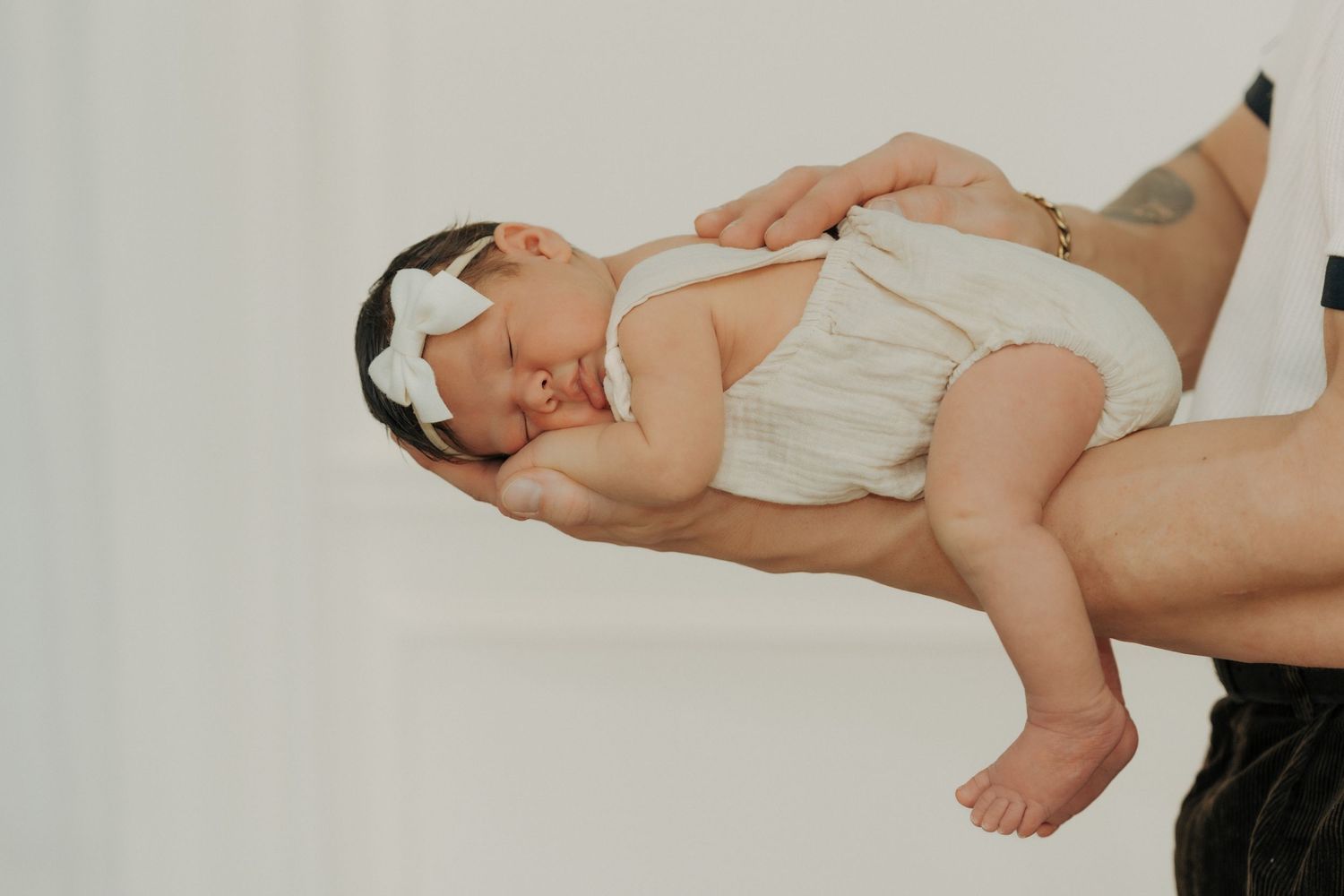 A baby wearing a white onesie lies sleeping peacefully on a parent's lap against a white background.