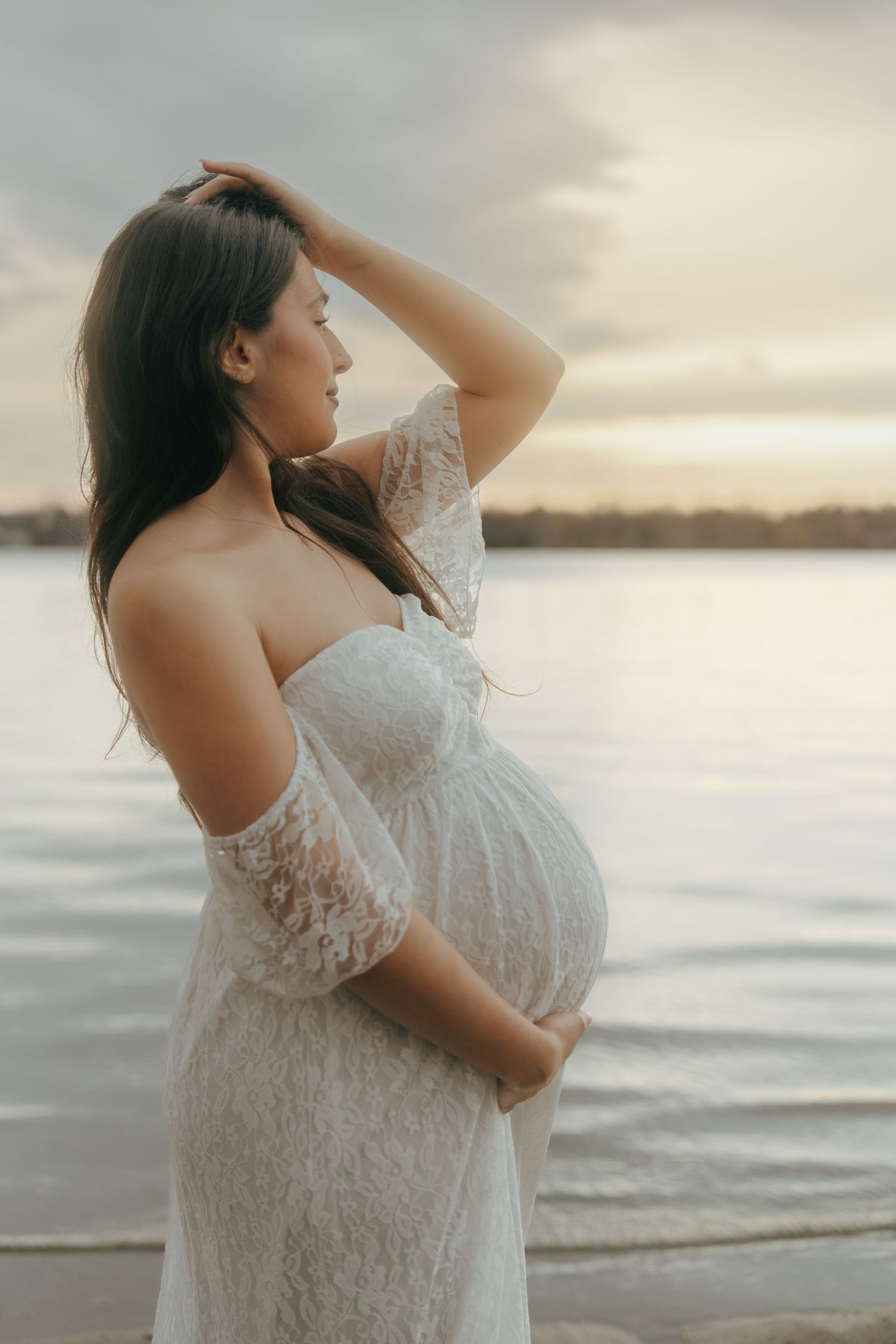 A pregnant woman in a white lace dress poses at sunset by the water in a dreamy maternity photoshoot.