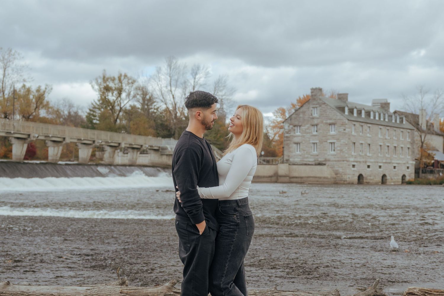 A couple shares a romantic moment on a rocky beach with historic buildings in the background during autumn.