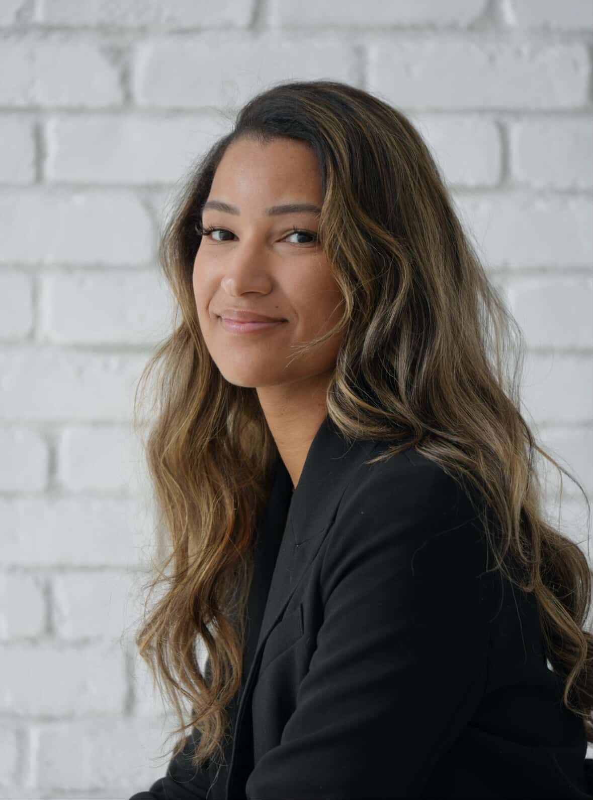 Professional woman in dark blazer with wavy brown hair smiling against white brick wall background.
