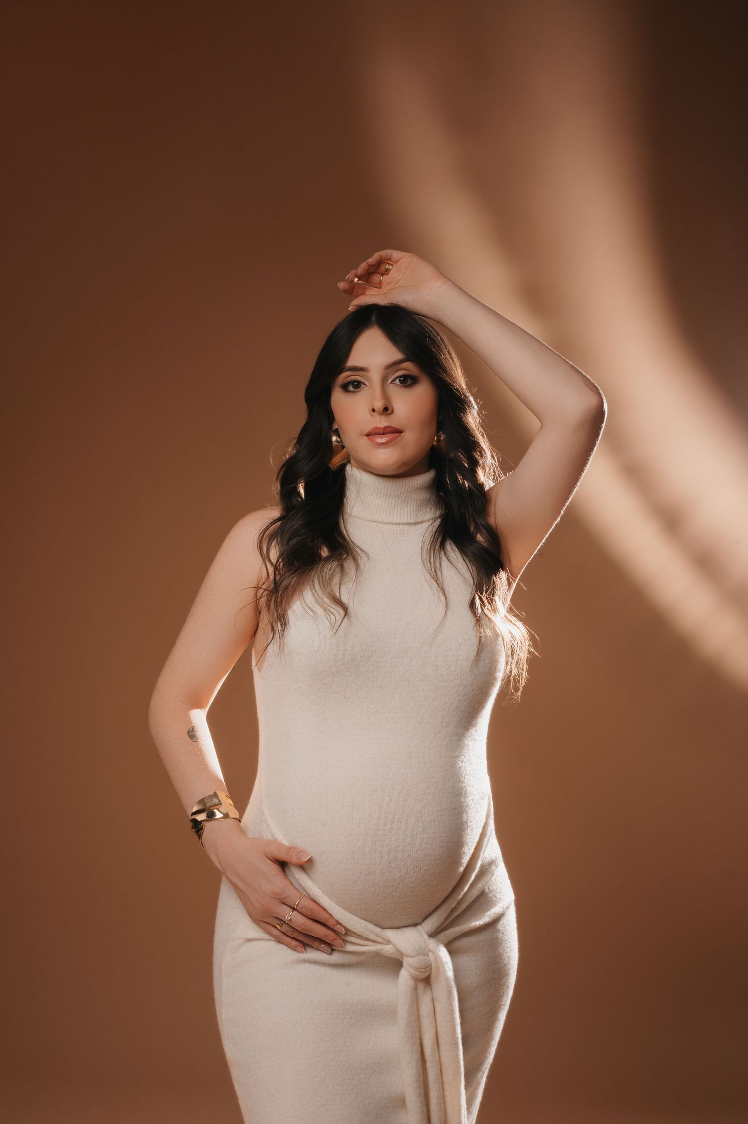 Retrato elegante de gestante de uma mulher em um vestido de gola alta cor creme posando contra um fundo marrom com iluminação dramática.