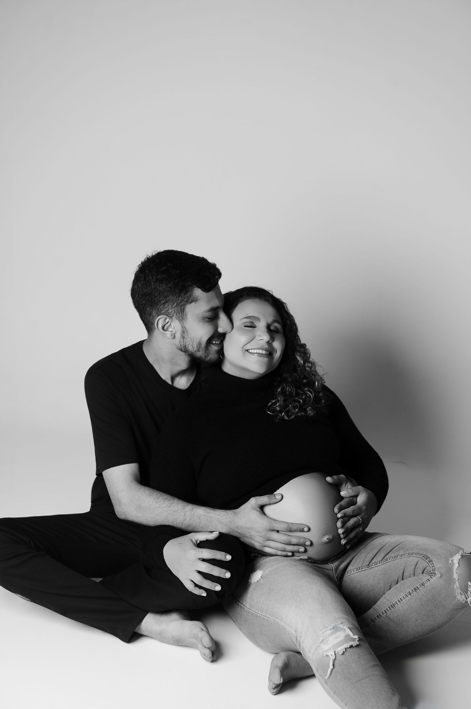 Ensaio de gestante em estúdio com o casal assentados no chão, juntos, felizes e de olhos fechados