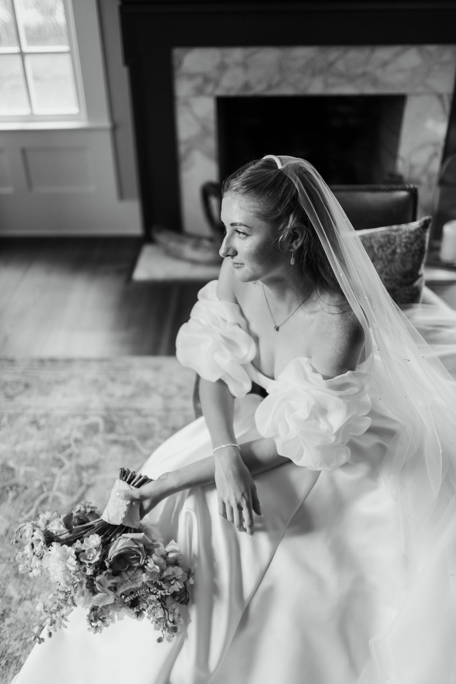 Black and white series of a bride in an elegant white gown with ruffled details sitting by a window with her bouquet.