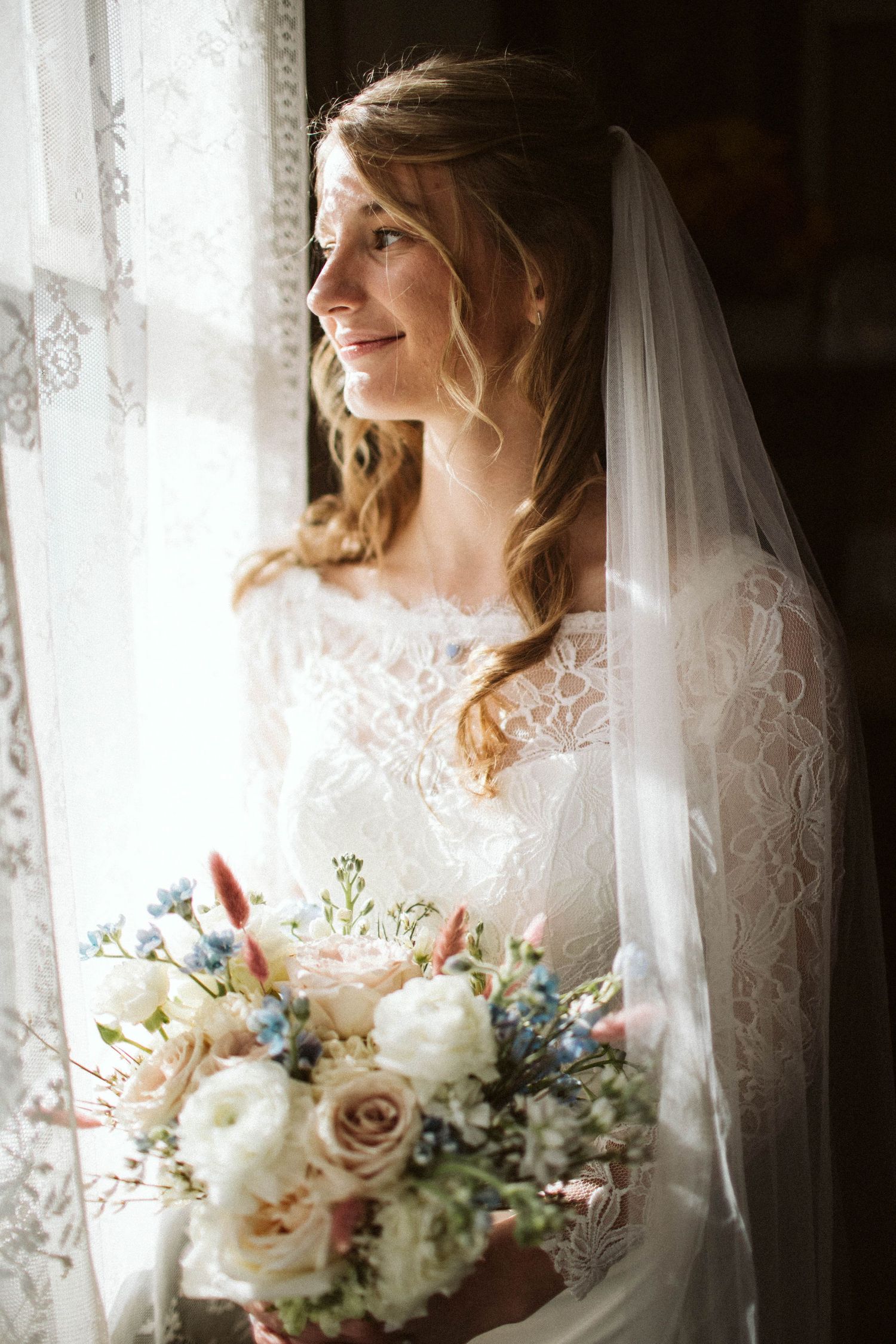 A bride in a white lace dress and veil stands by a window holding a pastel bouquet with roses and white flowers.