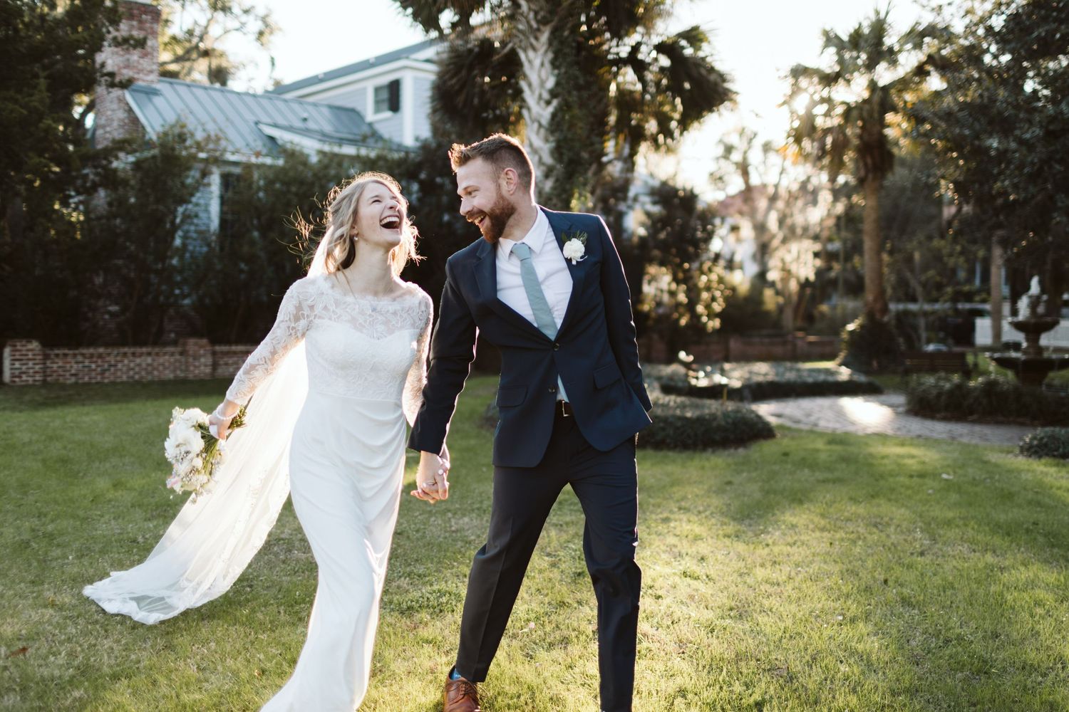 A couple in wedding attire embraces while standing in a sunlit garden setting at golden hour.