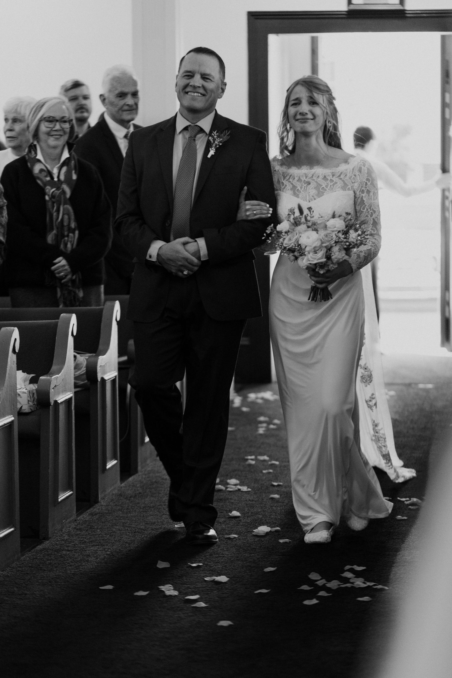 Black and white photo of a wedding processional walking down the aisle with rose petals scattered on the ground.