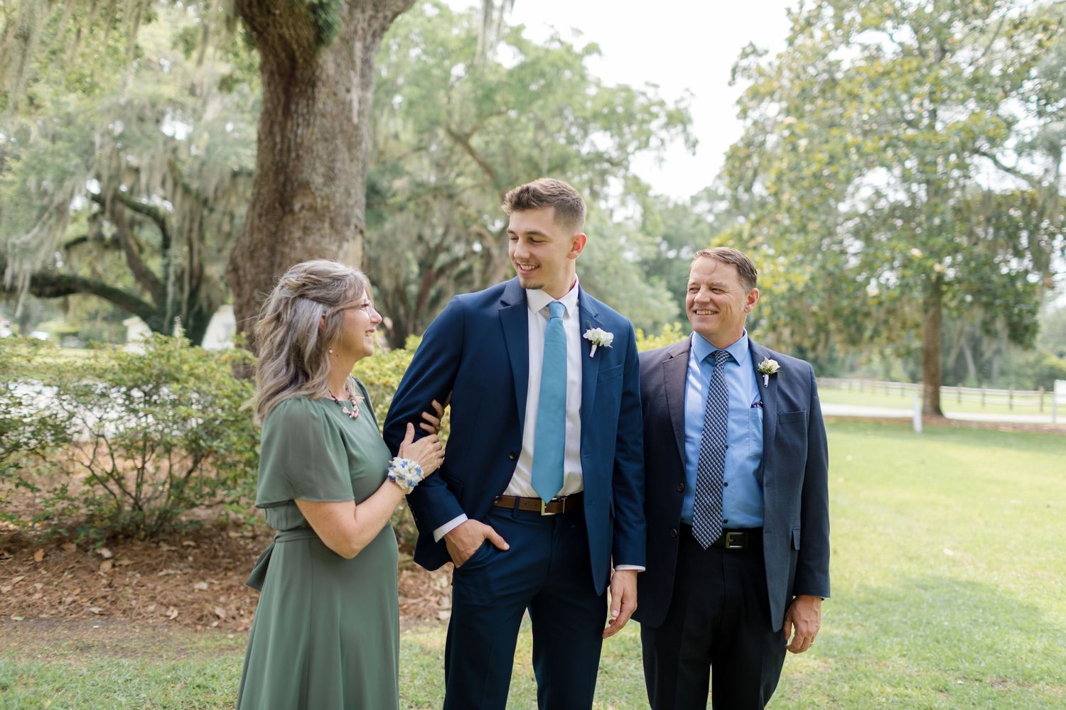 Wedding party members in formal attire laugh together while standing on grass in an outdoor garden setting.