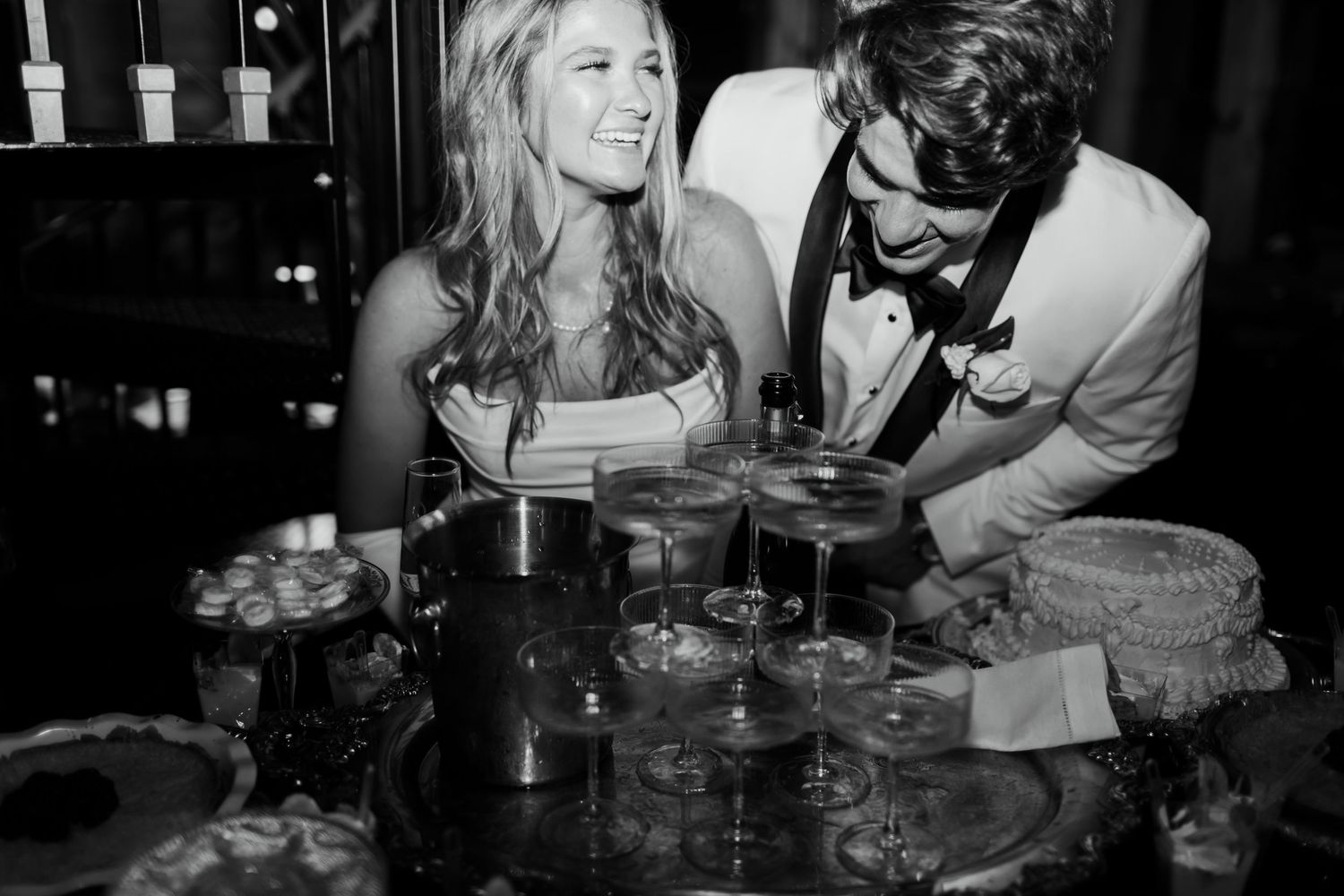 A black and white photo of a couple laughing together at a wedding reception while pouring champagne and cake.