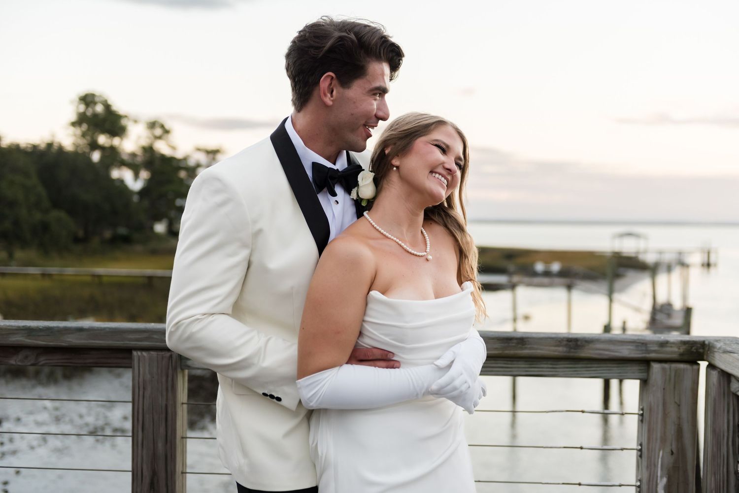 A romantic couple in formal attire embraces on a wooden dock during a low country sunset by the water.