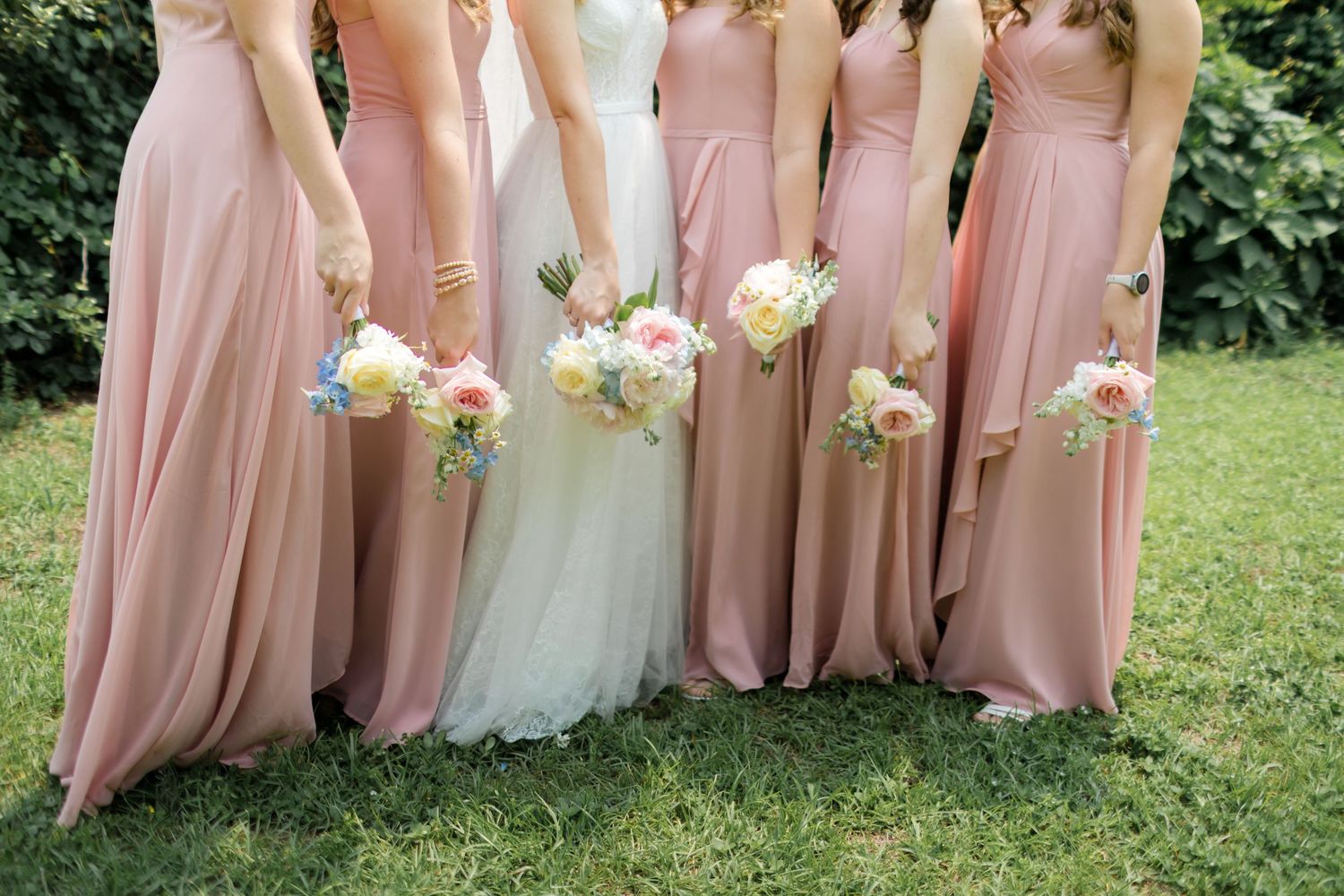A wedding party in blush pink bridesmaid dresses and a white bridal gown stand together on green grass outdoors.