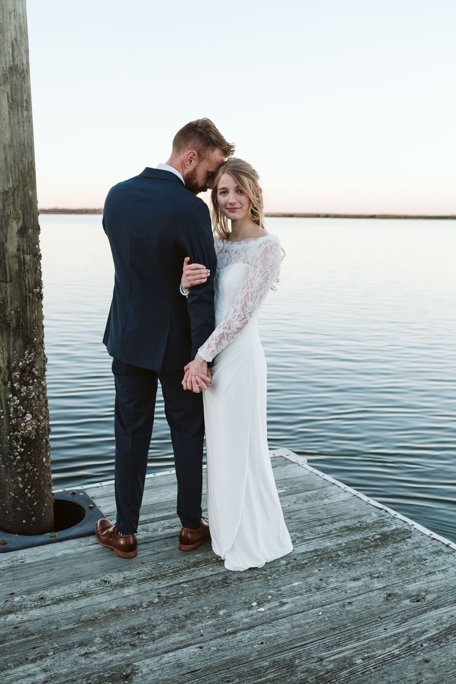 A couple in wedding attire shares an intimate moment on a wooden pier overlooking a serene waterfront at sunset.