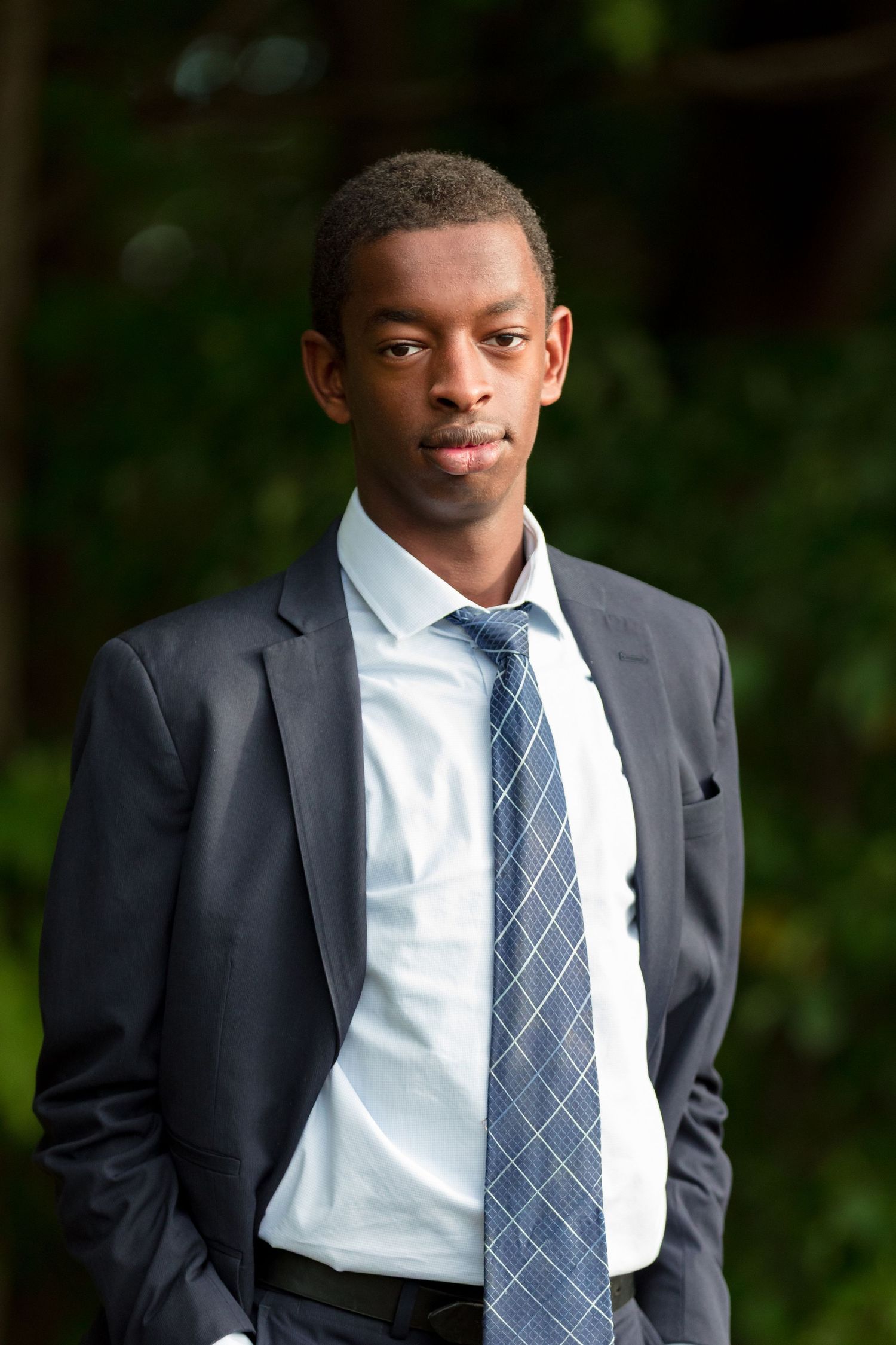 A person in formal attire poses against a natural green background.