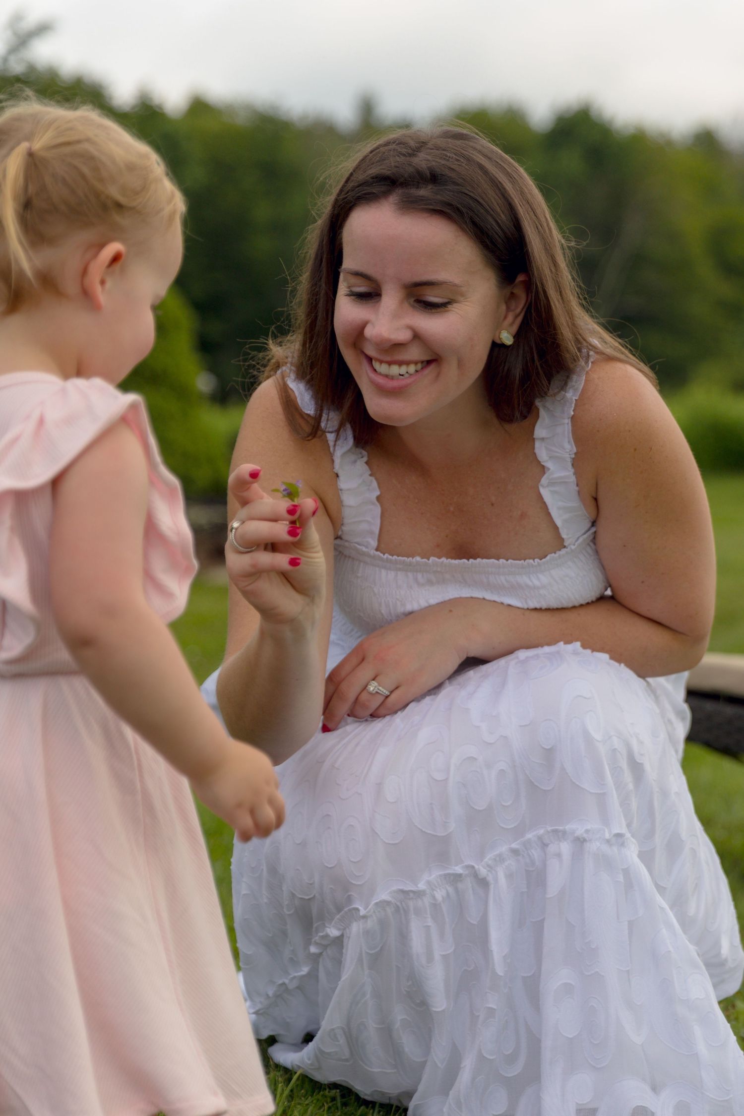 A woman in a white dress sits on grass while interacting with a child in a pink outfit.