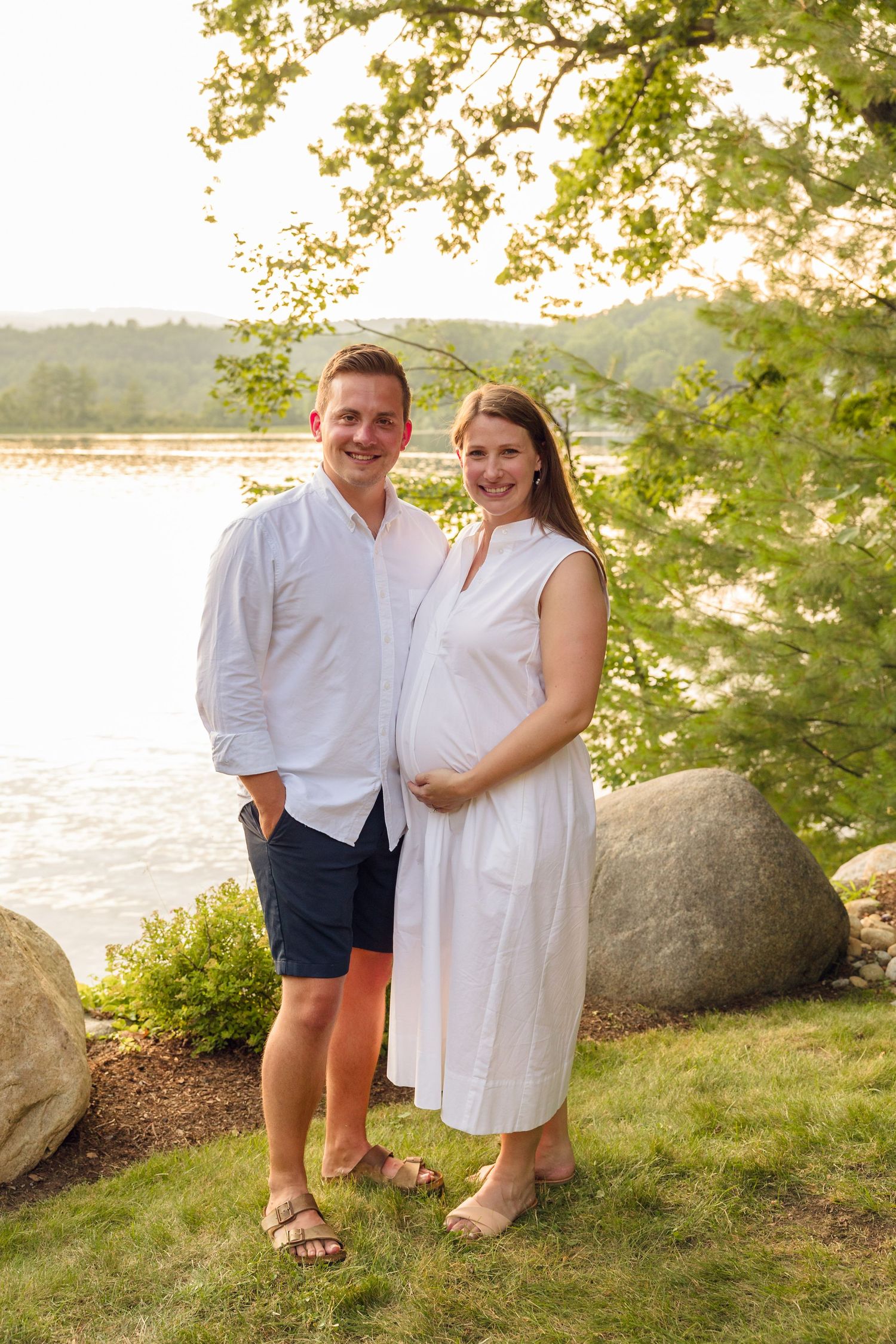 Couple in white summer outfits poses together near a lakeside with large rocks.