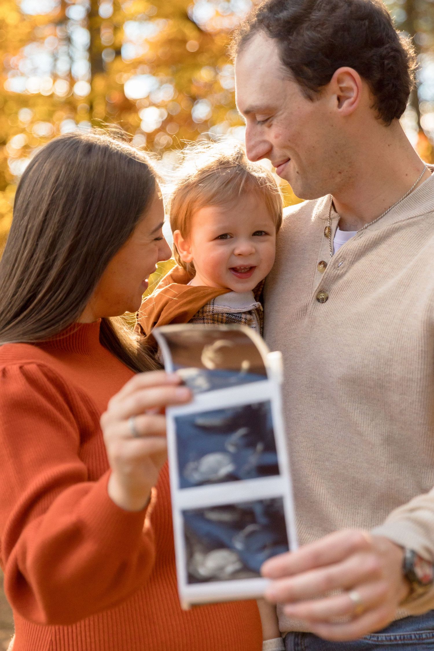 Family shares joyful moments while looking at ultrasound photos in golden autumn sunlight.