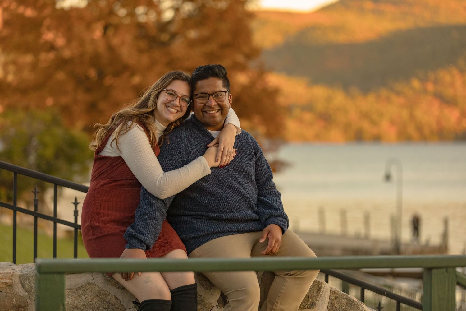 Couple embracing on lakeside deck during golden hour with autumn colors in background.
