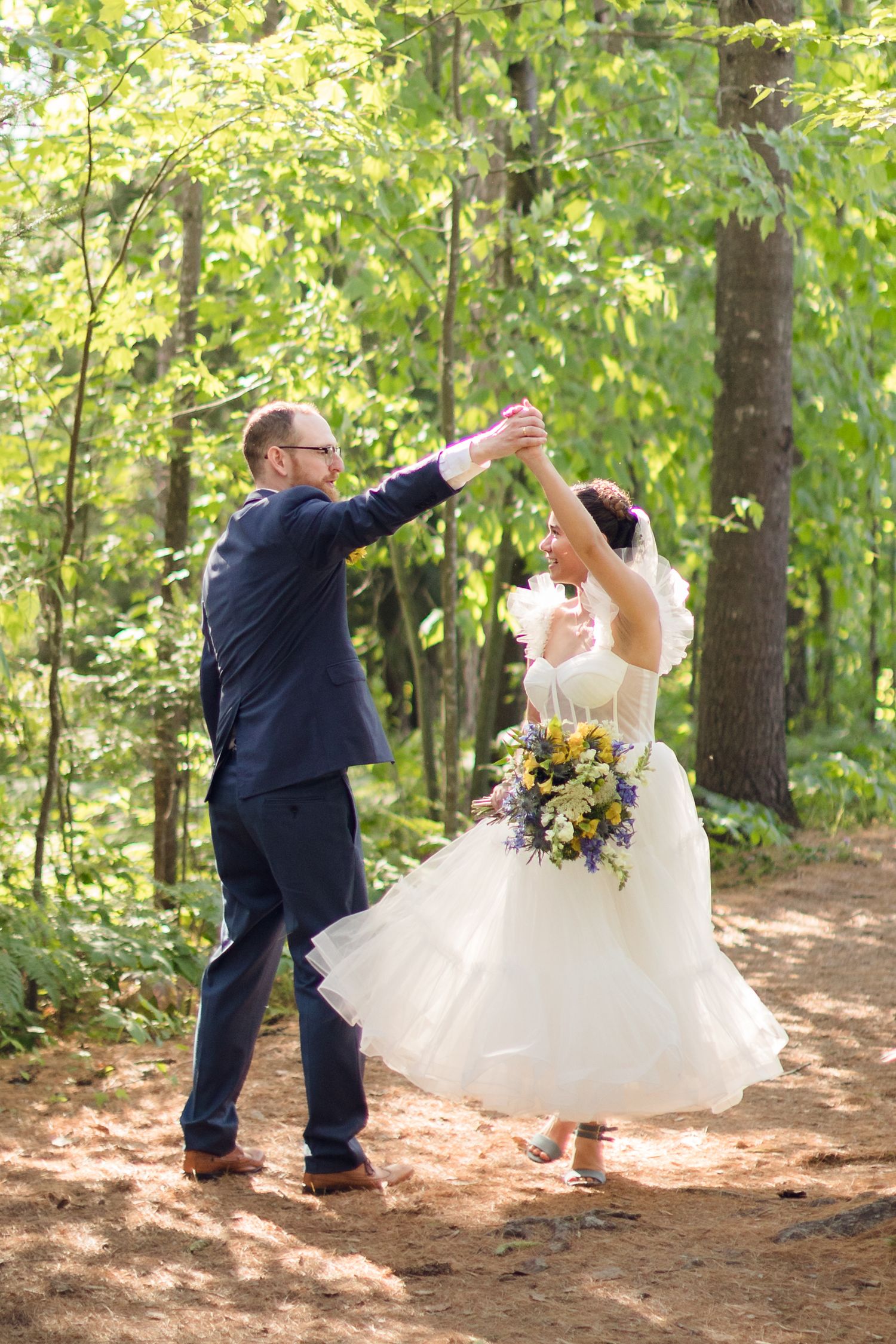 A couple in wedding attire dances playfully in a wooded forest setting with sunlight filtering through green leaves.