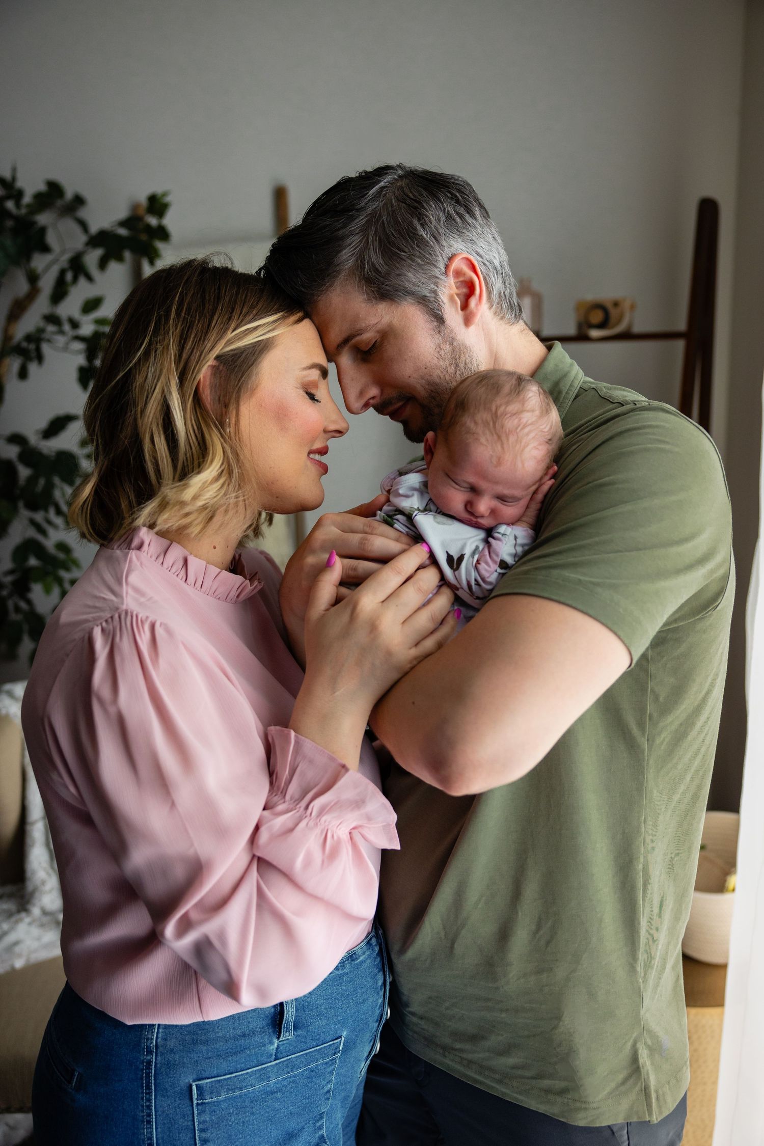 A couple in pink and green shirts share an intimate moment while holding their newborn baby at home.
