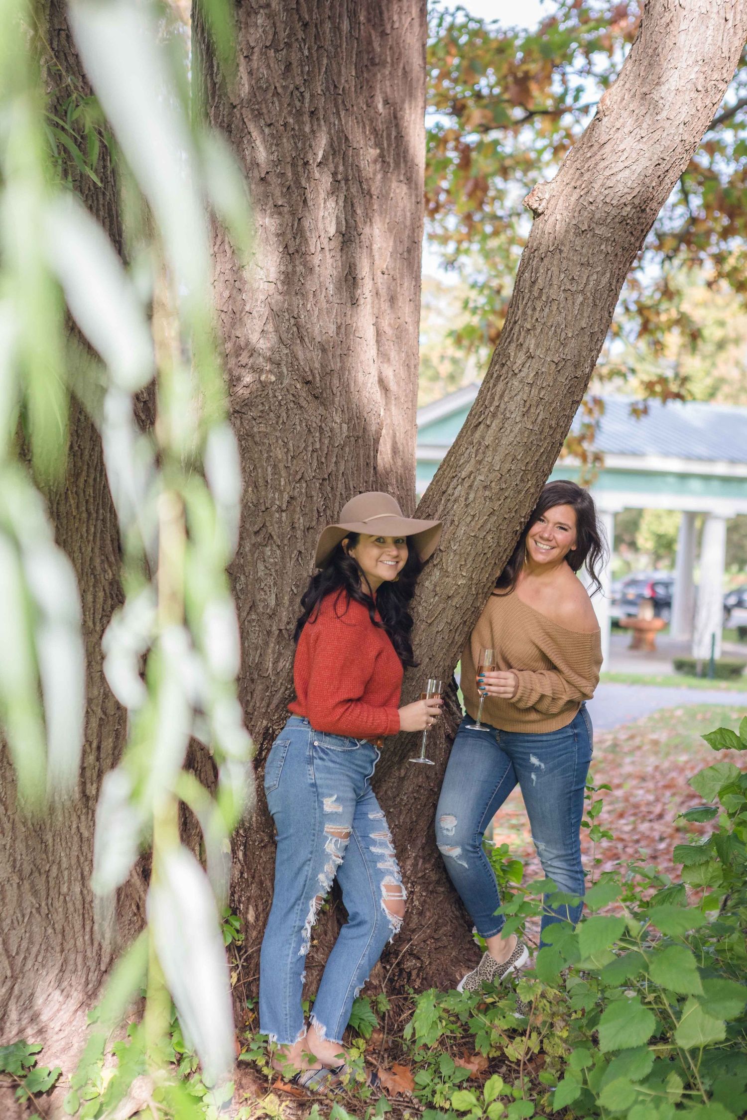 Two women in jeans leaning against a large tree outdoors, one wearing a cowboy hat, holding wine glasses.