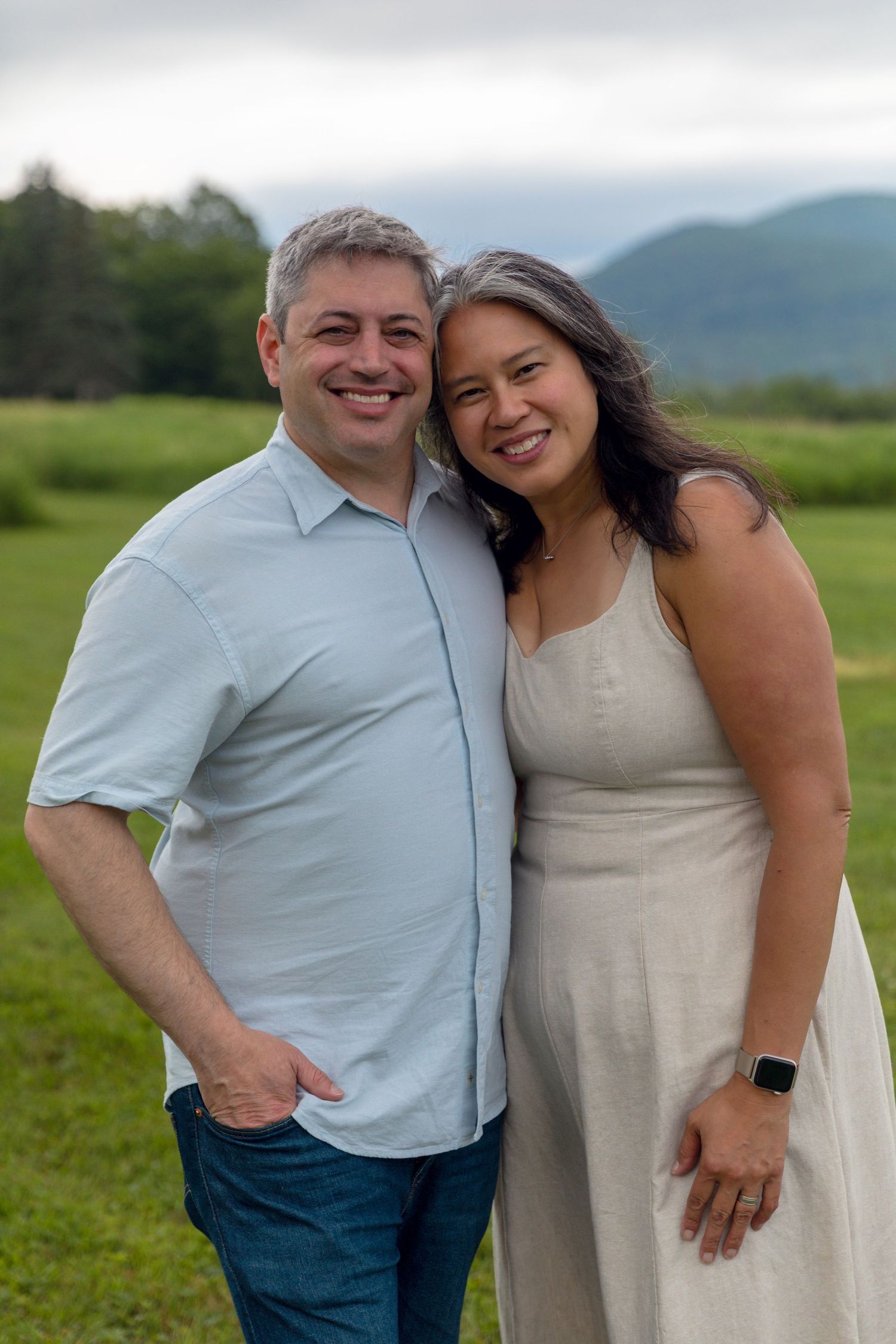 A couple embraces while standing in a green field with mountains visible in the distance.