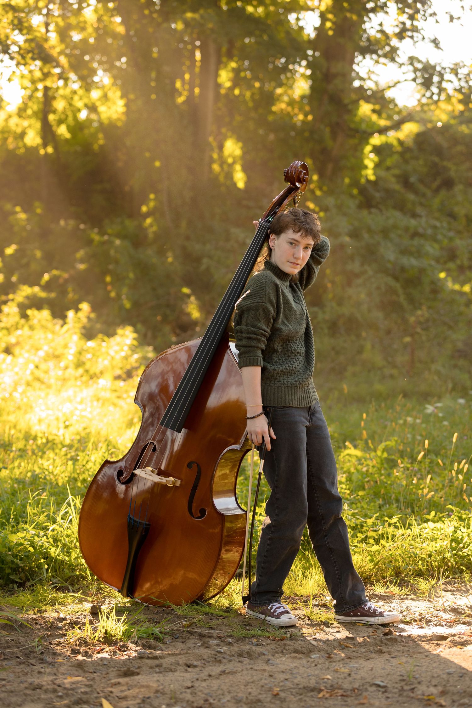 Musician poses with upright bass in sunlit forest clearing at golden hour.