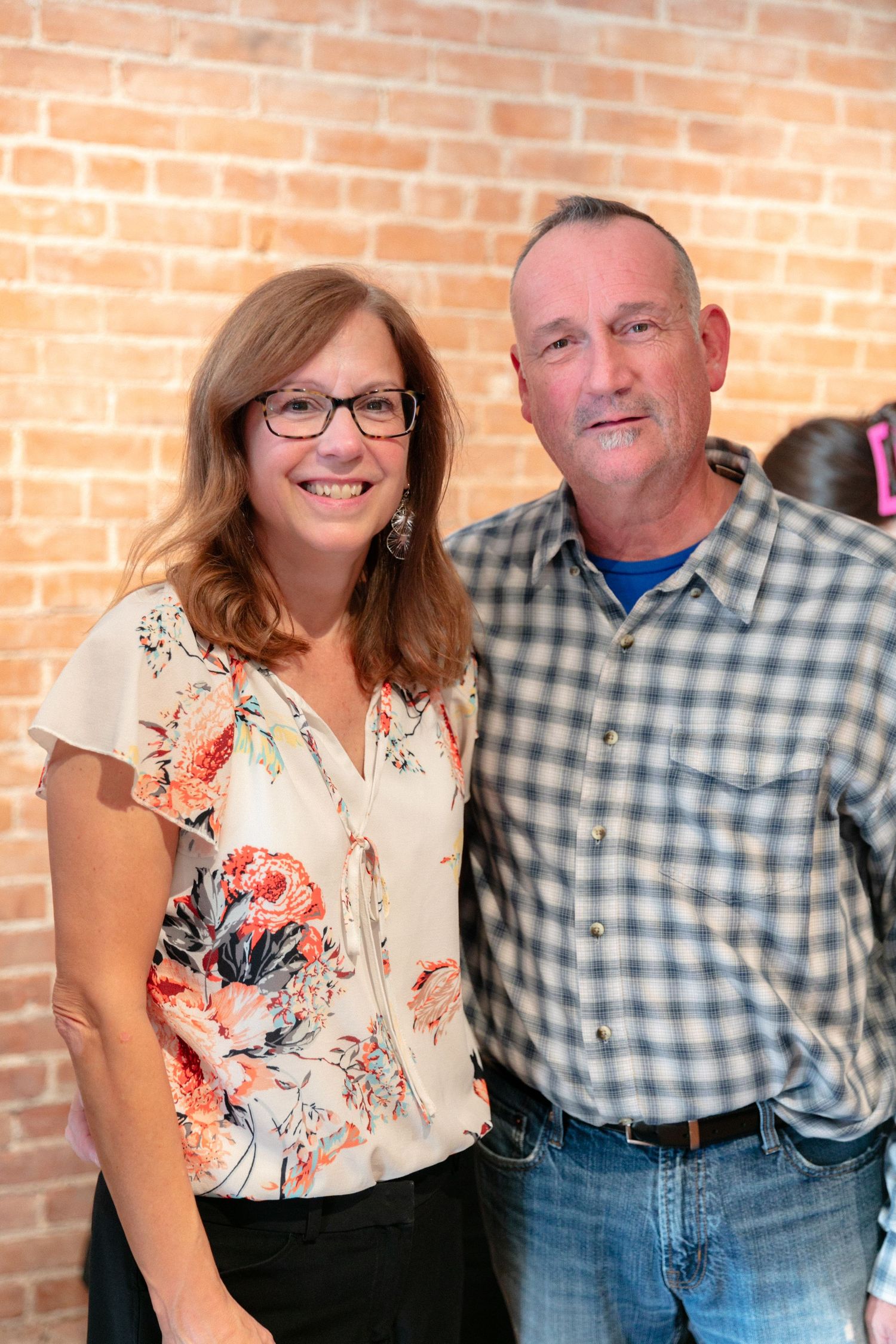 Couple poses together against brick wall, woman in floral top and man in plaid shirt.