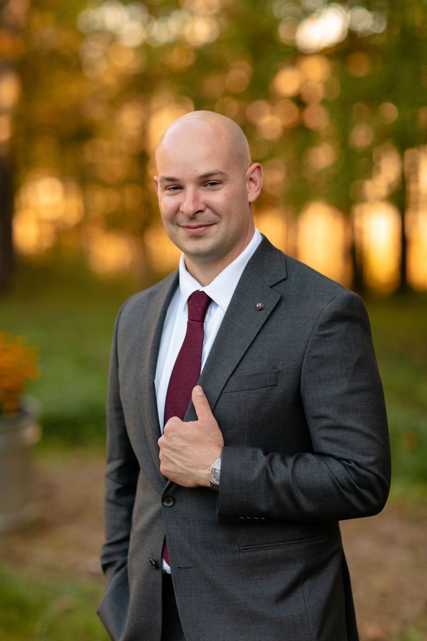 Groom in charcoal suit adjusts his burgundy tie while smiling in an autumn outdoor setting.