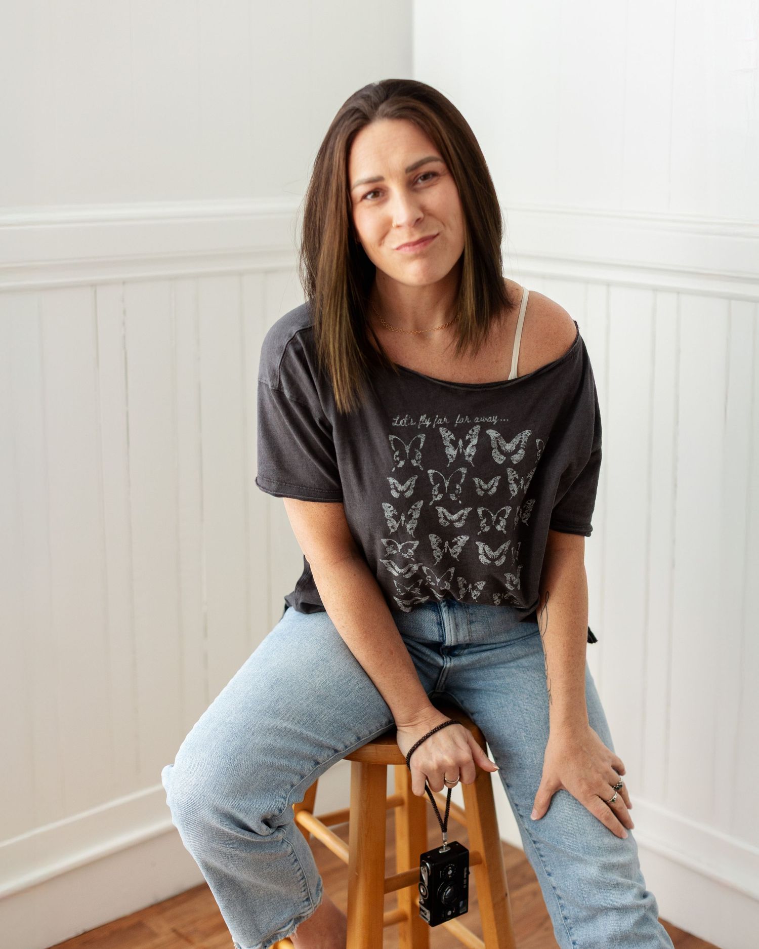 Person in casual attire poses on wooden stool against white wall with natural lighting.