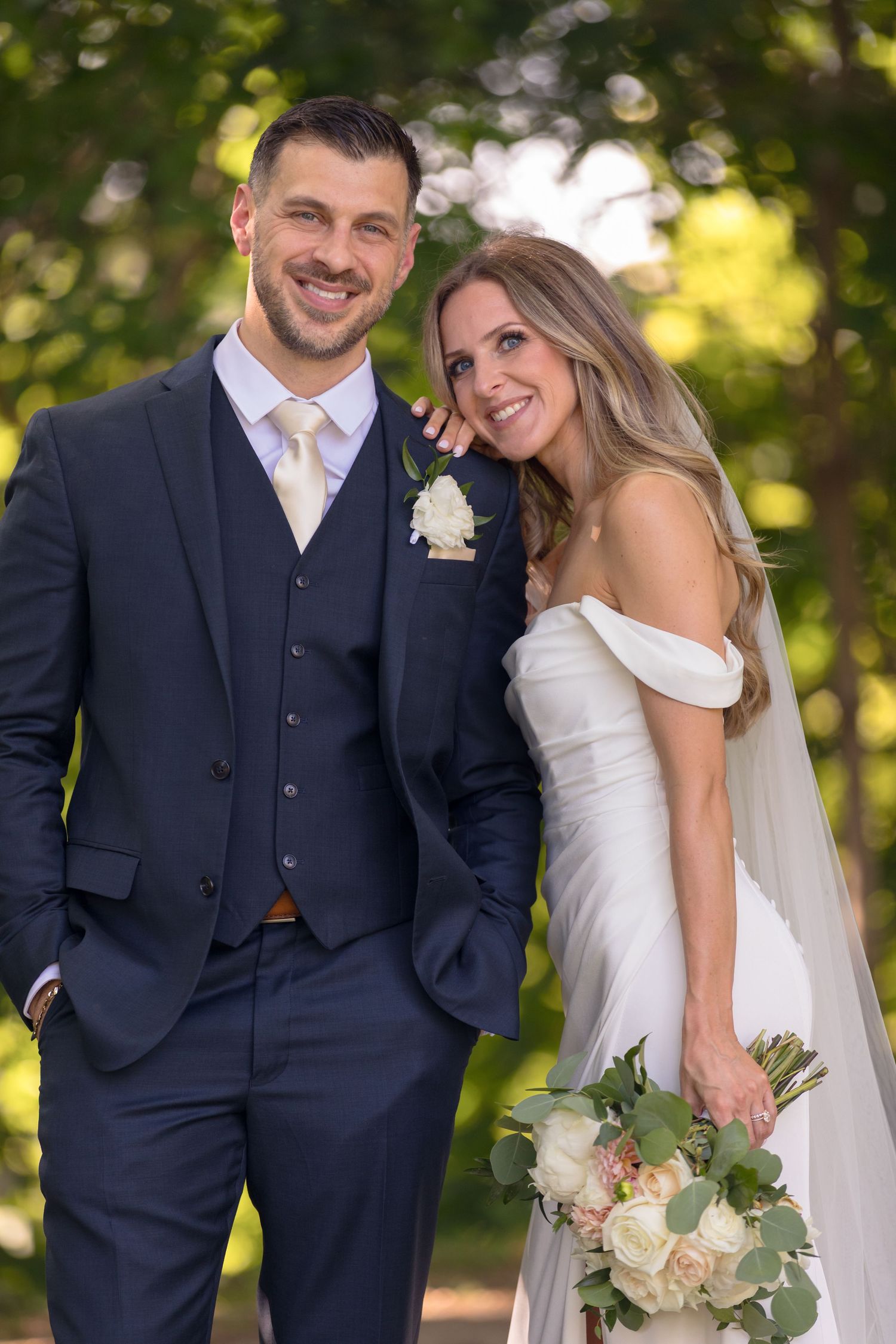 A newlywed couple poses outdoors on their wedding day, with the bride in white and groom in navy blue suit.