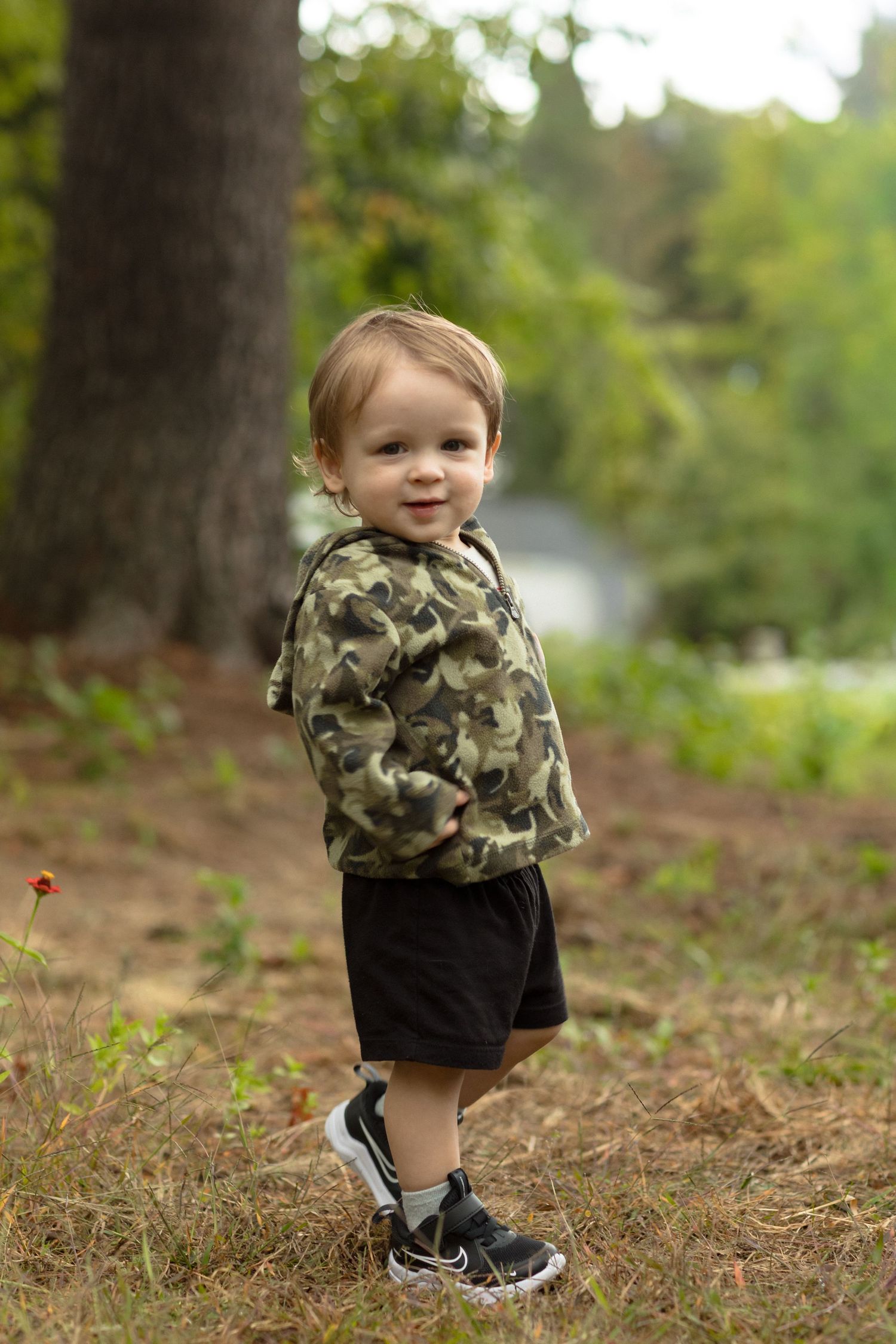 A small child wearing camouflage and black shorts plays outdoors in a wooded area.