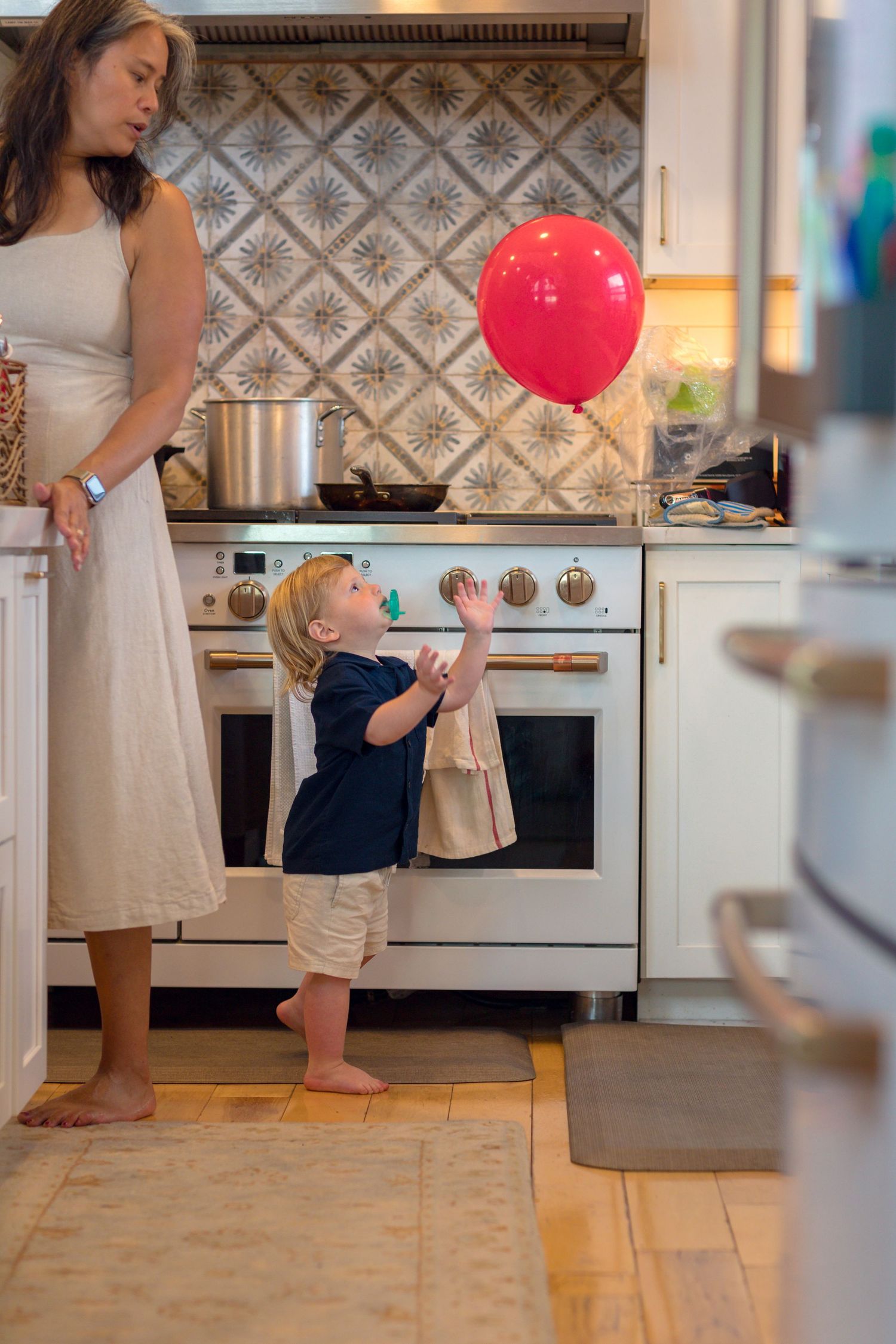 A person in a beige dress interacts with a child near a red balloon in a kitchen with decorative tile backsplash.