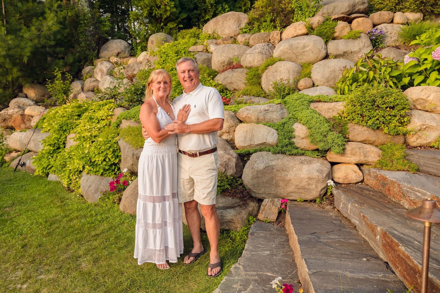 A couple stands together on a stone patio with a landscaped rock garden wall in the background at sunset.