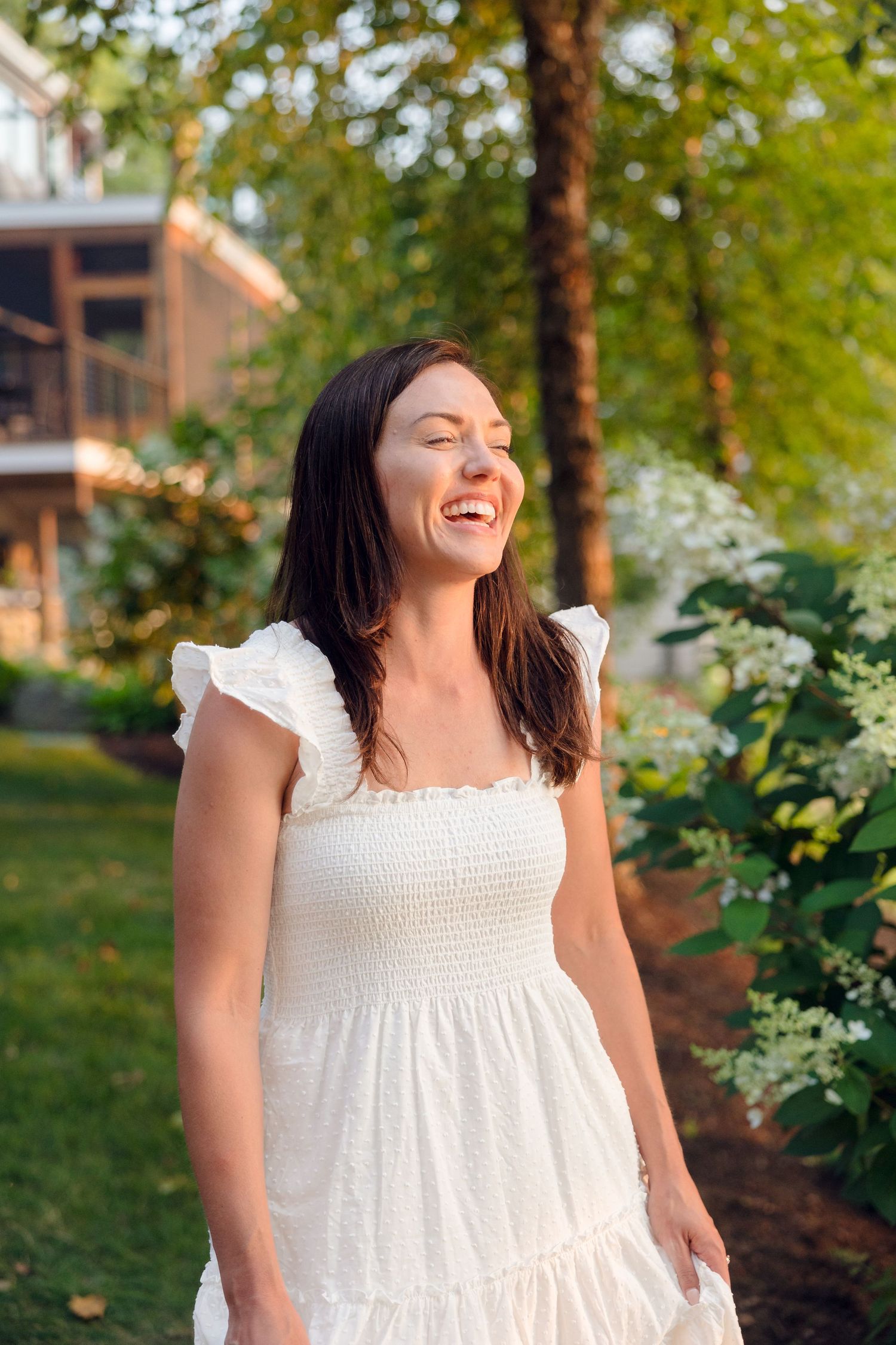 A woman in a white summer dress twirling and laughing in a garden setting.
