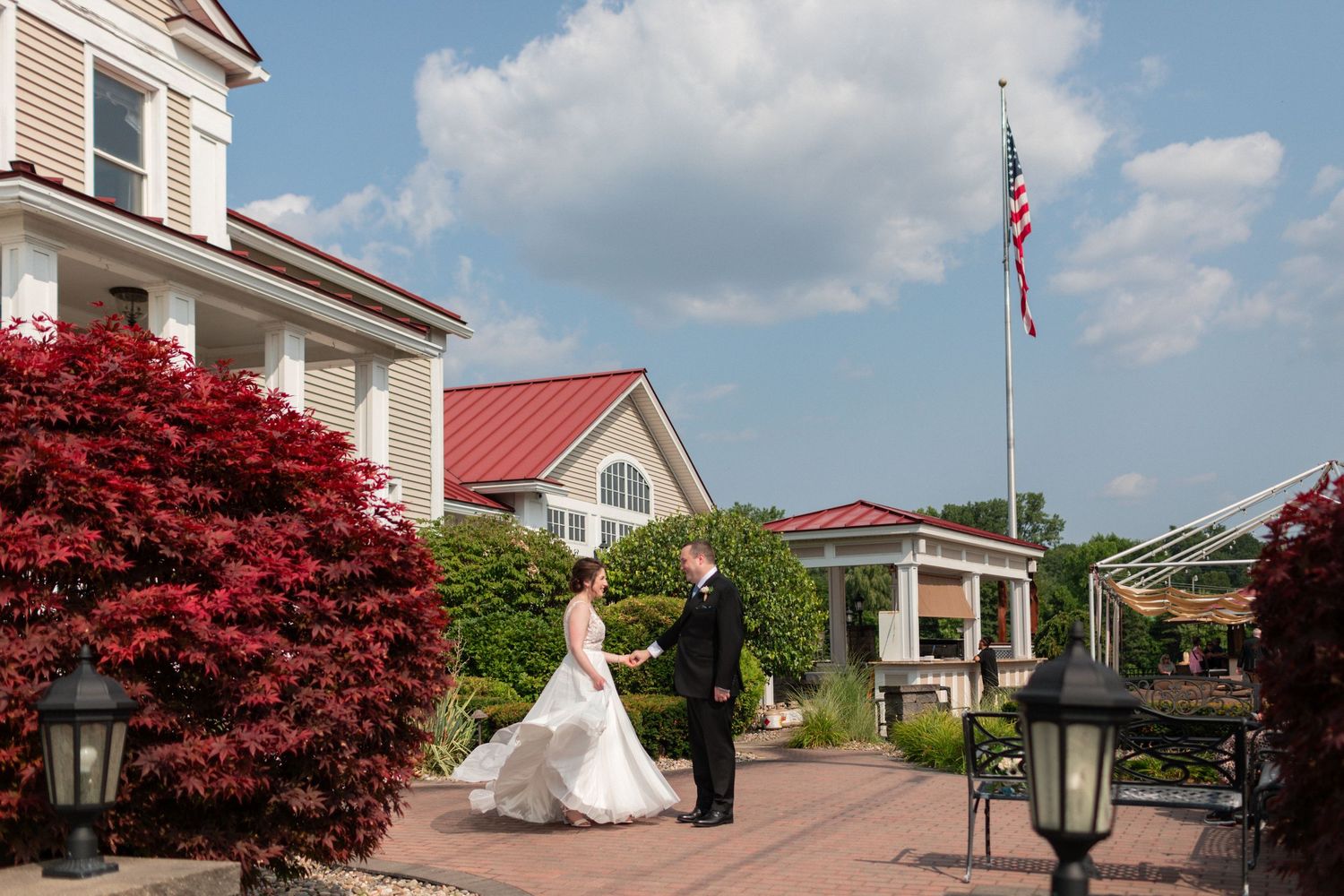 A wedding couple stands on a brick courtyard with an American flag and red maple tree in view.